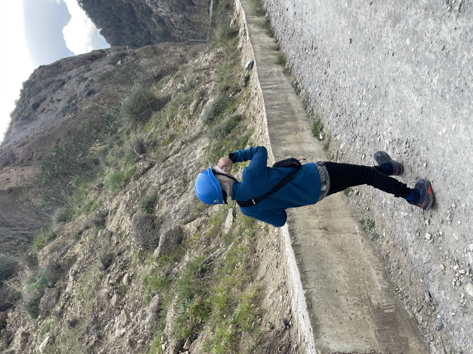 The image, when viewed in its correct orientation, captures a person standing on a rugged path amidst a mountainous landscape in El Chorro, Spain. The scene unfolds during daytime, under a bright, partly cloudy sky.
The central figure is an individual, likely an adult, attired in functional outdoor clothing. They wear a blue long-sleeved top, black pants, and athletic footwear, with one shoe appearing blue/black and the other red/black. A prominent blue hard hat or helmet protects their head, featuring a white logo, which appears to be "PETZL," indicating climbing or safety gear. A black fanny pack or sling bag is worn across their chest.
The person is shown in a three-quarter back view, looking upwards and towards the right side of the frame, seemingly observing the imposing, rocky cliffs that form the backdrop. Their right hand is raised near their face, possibly adjusting the helmet, shading their eyes from the sun, or holding a small item.
The immediate foreground includes a narrow, unpaved dirt path to the right, adjacent to a weathered concrete channel that stretches into the distance. This concrete structure, possibly an old aqueduct or part of a constructed trail, runs parallel to the path. To the left of the channel, the terrain rises steeply, characterized by loose rock scree, interspersed with patches of sparse green grass and small, resilient shrubs. The environment suggests a natural, arid, and challenging outdoor setting, typical of the dramatic gorges and climbing areas found in El Chorro.