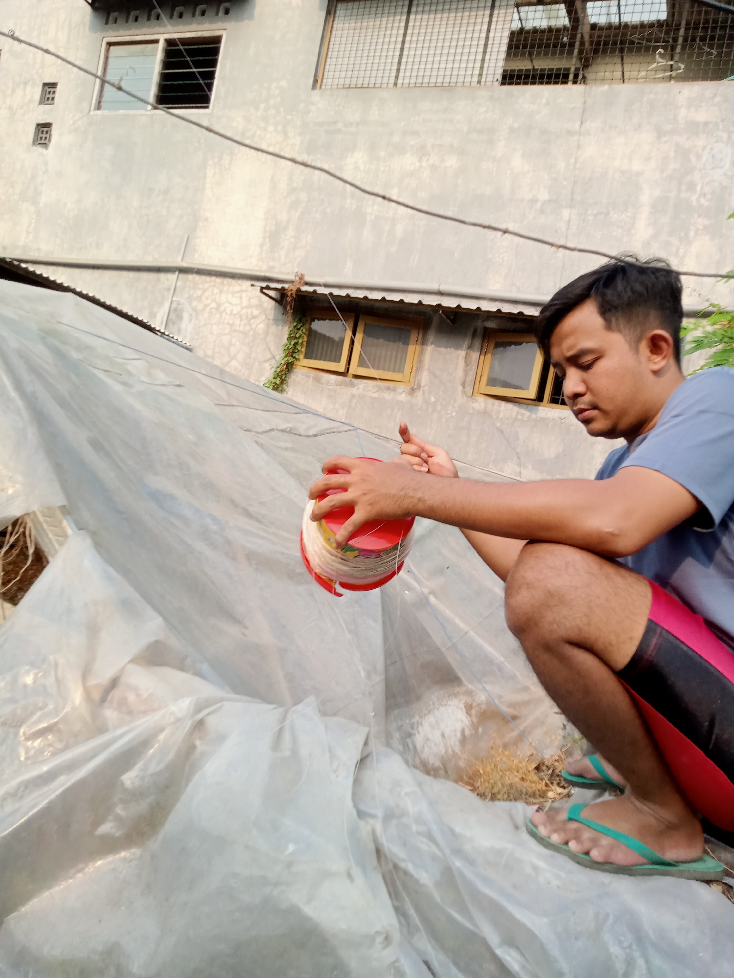 A man is squatting next to a large structure covered in translucent plastic sheeting. He is holding a red container with a spool of white string. The man is wearing a blue t-shirt and red and black shorts. He has green flip-flops on his feet. The background shows a building with windows and an overcast sky. The location is Simo, Indonesia.