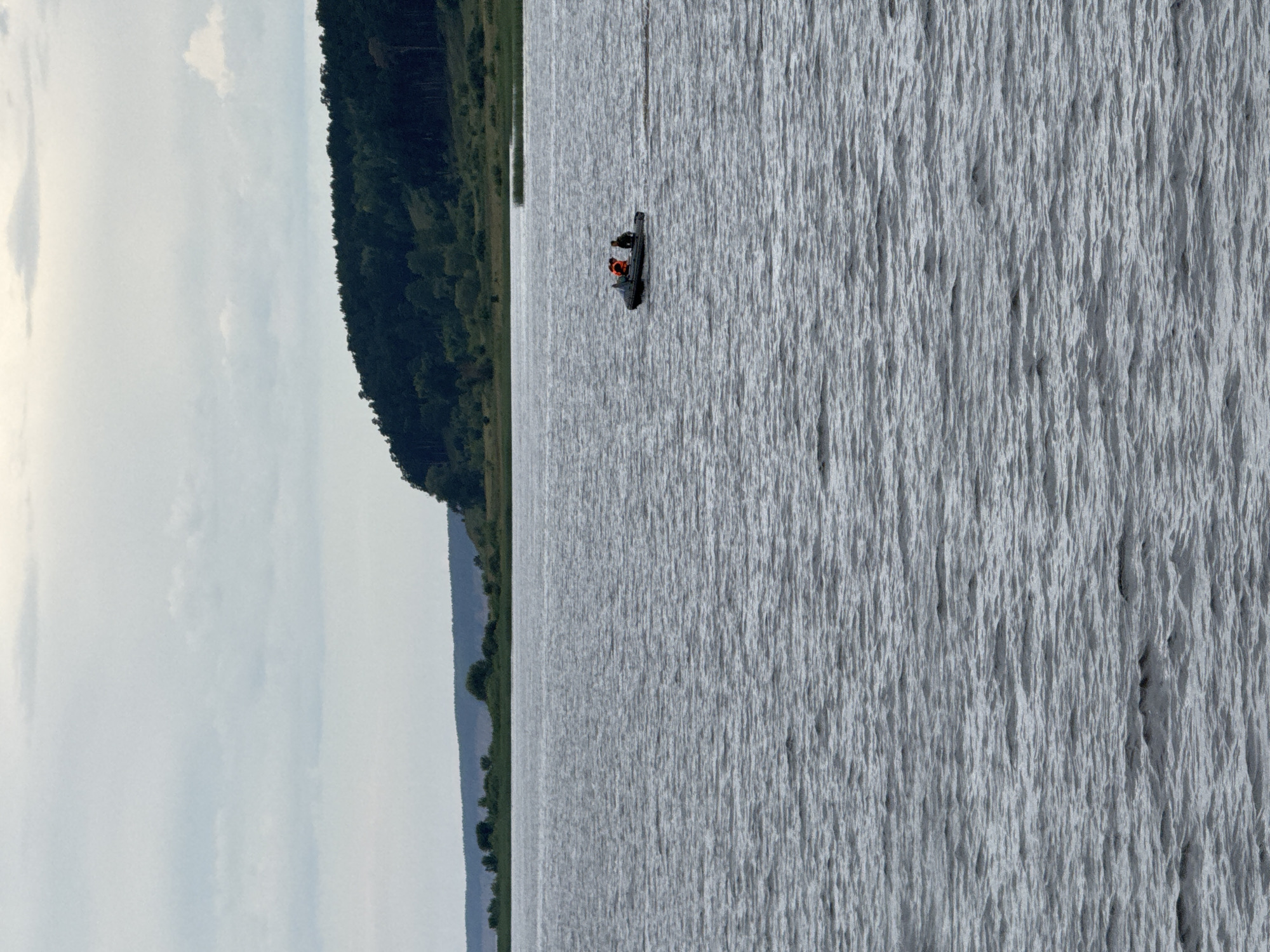 An eye-level, medium shot captures a wide, rippling body of water under a cloudy sky. In the middle distance, a small, dark inflatable boat with two occupants is moving across the water, leaving a slight wake. One person in the boat wears an orange life vest. The background features a tree-lined hillside with a darker band of forest along its top, and a strip of lighter green vegetation at its base, bordering the water. A hazy, light-colored band of land is visible further in the distance. The scene appears to be a natural landscape, likely a lake or a wide river, with no visible urban elements or specific landmarks indicating the city of Красный Бор, Russia.