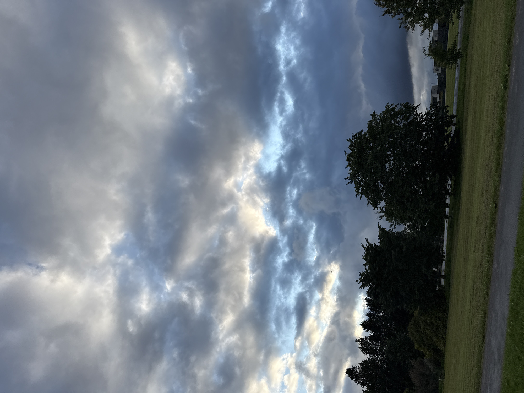 The image captures a view of the sky and a grassy field with trees in Järveküla, Estonia. The sky is dramatic, filled with a mix of dark, heavy clouds and patches of lighter, brighter clouds, suggesting a transition in weather or an approaching storm. Sunlight breaks through in some areas, creating a luminous effect against the darker clouds. In the foreground, a green lawn is visible, with a paved path running along the bottom. A line of dark green trees forms a backdrop to the field, with some of the trees appearing dense and full. In the distance, faint outlines of buildings can be seen through the trees and under the moody sky. There are no visible people or animals in the scene.