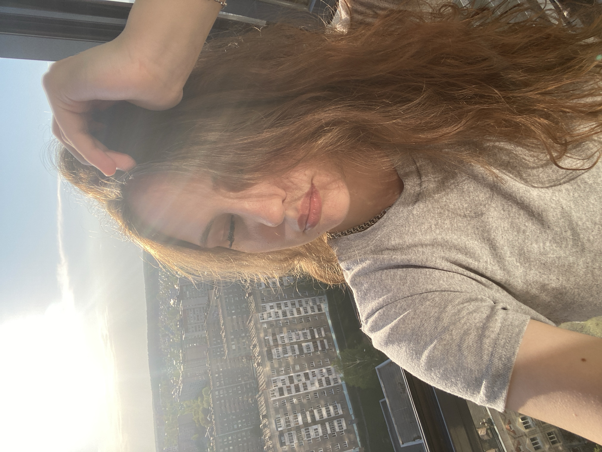 A person with long, light brown hair and wearing a gray t-shirt is taking a selfie. The person's left hand is touching their hair. The background shows a cityscape with numerous buildings and green spaces. The sunlight is bright, causing glare on the left side of the image. The location is Izhevsk, Russia, as indicated by the buildings.