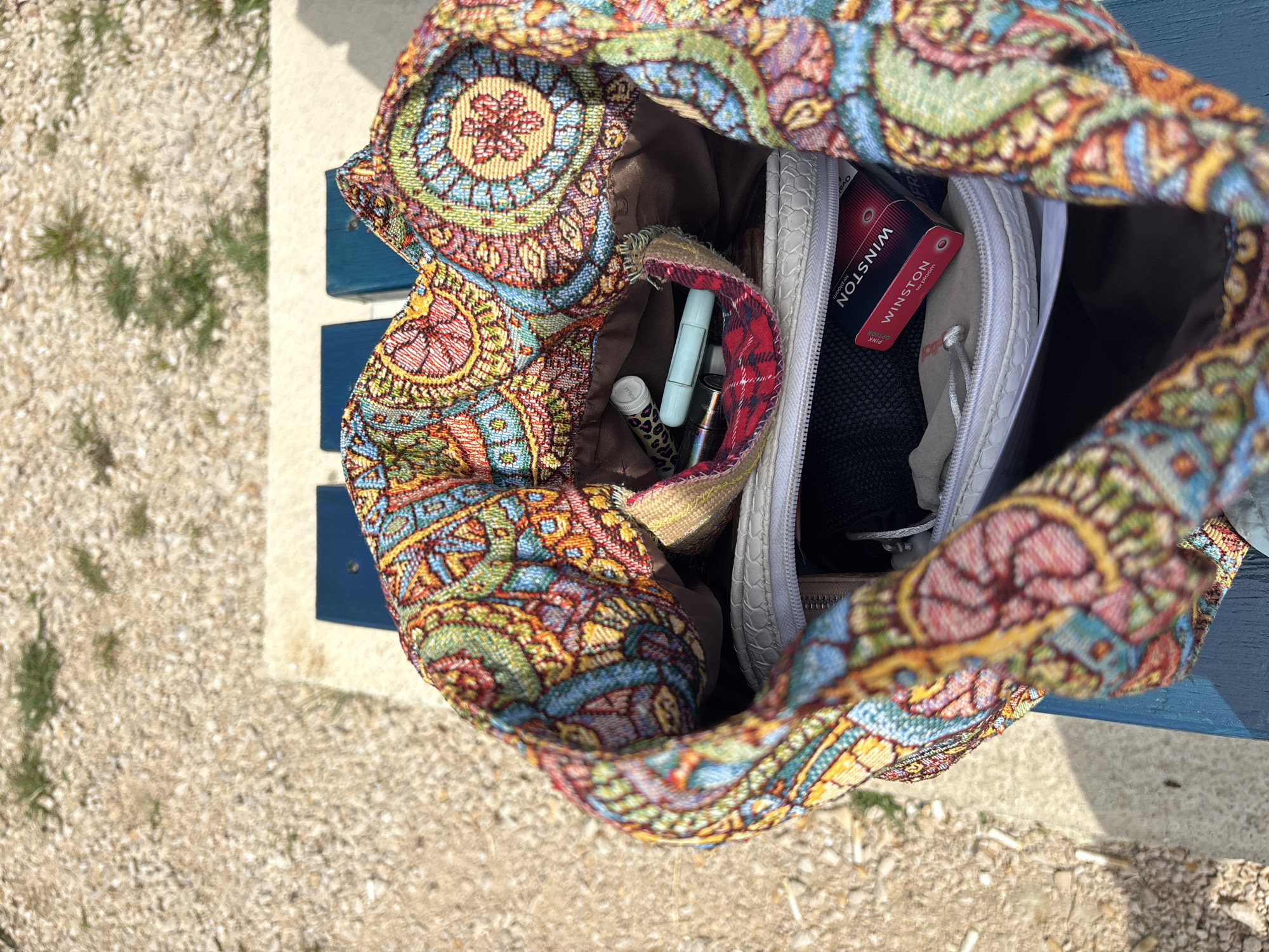 An overhead, close-up shot captures the open top of a brightly patterned fabric bag, revealing some of its contents. The bag features an intricate, tapestry-like design with vibrant geometric and floral motifs in shades of blue, green, red, yellow, and orange. The fabric appears woven or embroidered. Its interior is lined with brown fabric, and a small internal pocket displays a red and black plaid pattern.
Inside the bag, several personal items are visible. On the left, a white tube (possibly lip balm), a light blue cylindrical object (perhaps a pen or a small vape), and a metallic patterned tube are seen. On the right, a white zippered pouch with a textured, possibly faux leather surface, holds further items. Within this pouch, a black mesh pocket is visible. A prominent red rectangular package is partially sticking out, clearly labeled "WINSTON" with "PINK OPTION" and "for cocoon" also readable. Another zippered compartment of the white pouch is also visible.
The bag is resting on a dark blue surface, which appears to be part of a wooden or metal bench. To the left and below the bench, the ground is covered in light-colored gravel with sparse patches of green grass, suggesting an outdoor setting. The bright, even lighting indicates it is daytime, likely sunny. No people or specific activities are depicted; the image focuses solely on the bag and its contents in Bar, Montenegro.