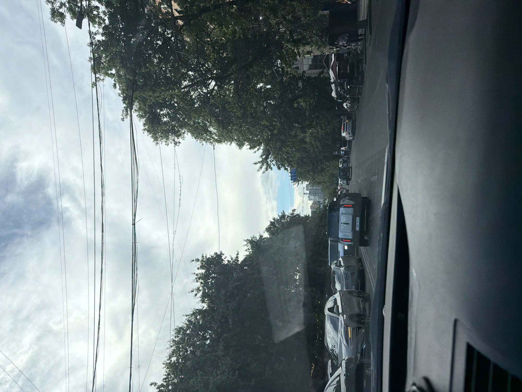 The image depicts a street view from inside a car in Yerevan, Armenia. The scene shows a traffic jam on a city street lined with trees and buildings. Several cars are visible in traffic, including a silver SUV and a dark colored vehicle with a license plate reading "338 TM 21." Overhead, power lines crisscross the sky. People are seen walking on the sidewalk and near parked cars. The sky is cloudy.