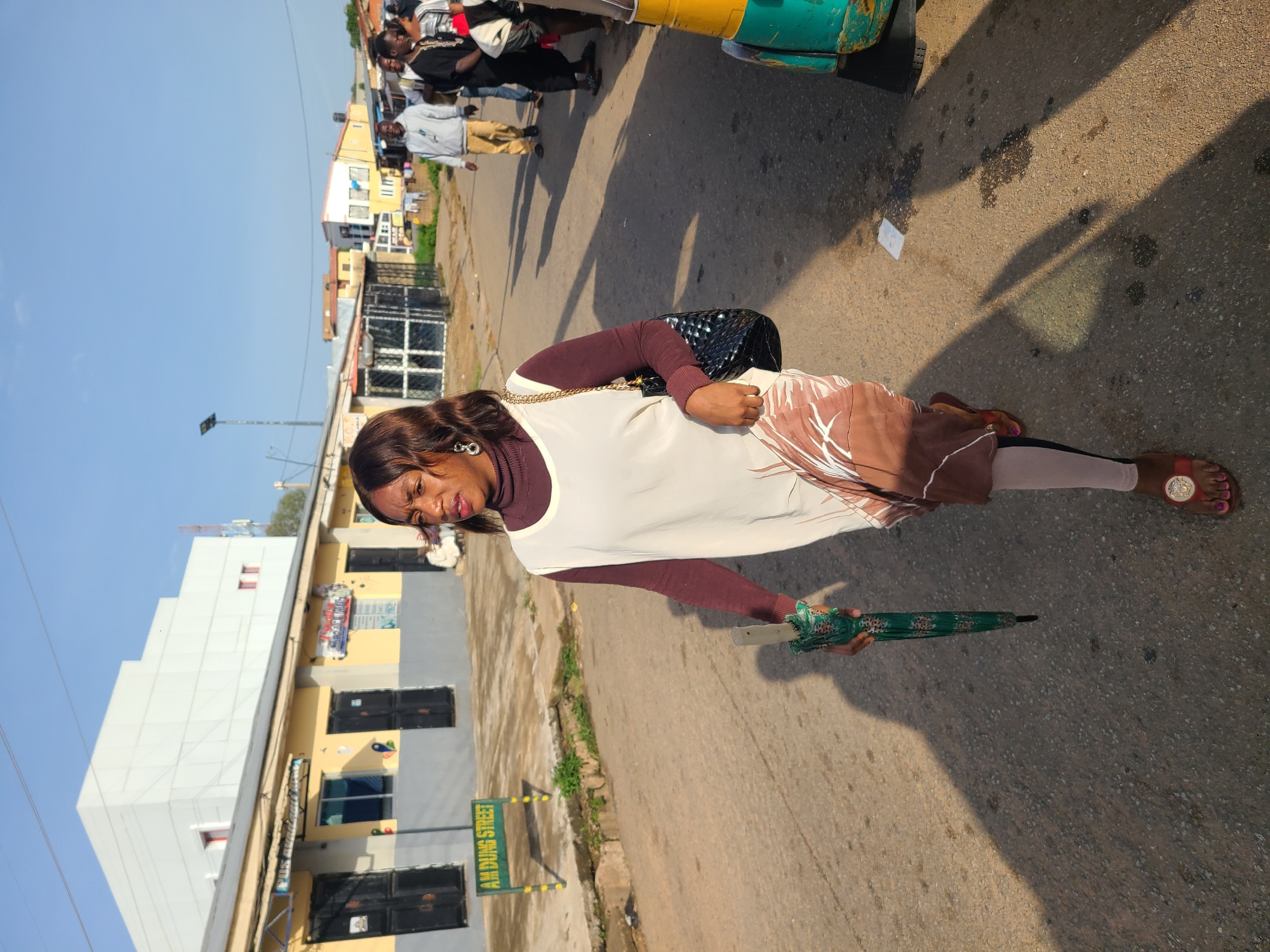 A medium shot captures a woman walking on a street in Jos, Nigeria. She is walking towards the right of the frame, holding a closed green umbrella in her right hand. She is wearing a brown turtleneck under a white sleeveless dress with a brown and white patterned hem. She has on light-colored leggings and brown sandals with gold embellishments. A black patterned handbag with a gold chain strap hangs from her left shoulder. Her expression is focused, and she is looking slightly to her left.
The street appears to be paved with asphalt and has patches of oil or water stains. The sun casts long shadows on the ground, indicating it is likely morning or late afternoon. To the left of the woman, a green and yellow sign reads "AM DUNG STREET". In the background, there are low-rise buildings with yellow facades and dark-colored doors and windows. One building is larger and white with red-framed windows. Other people are visible in the background, walking or standing on the street. A colorful motorized rickshaw, painted in green and yellow, is parked on the right side of the frame. The sky is clear and blue.
