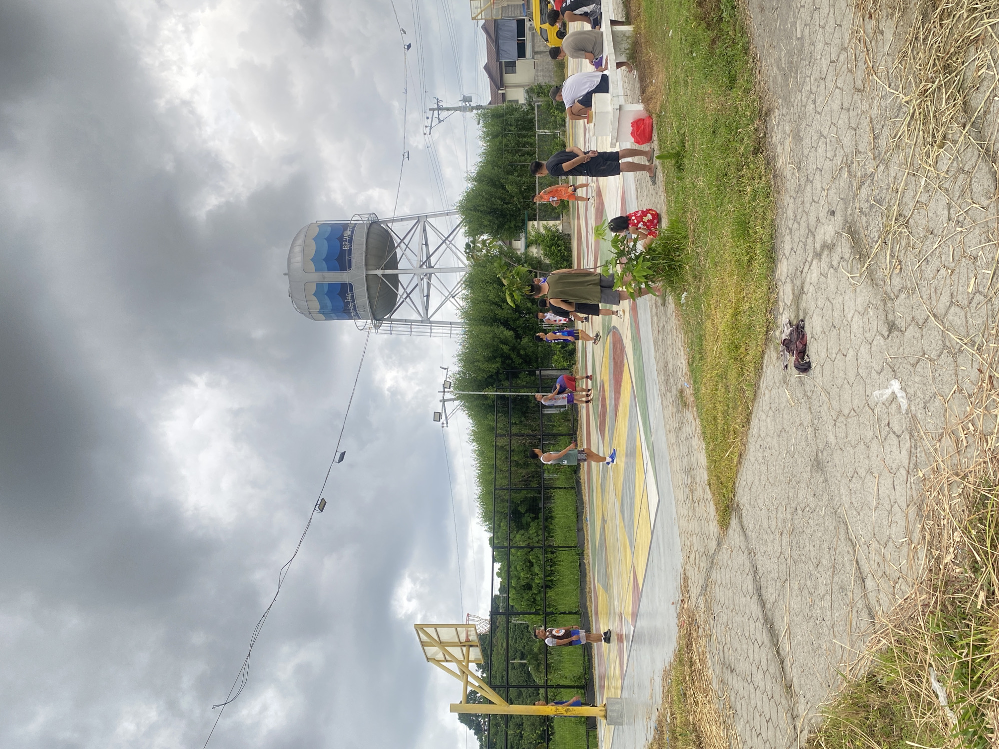 The image depicts a basketball court in Davao City, Philippines, with a towering water tank in the background. Several individuals are present on and around the court. Some are actively playing basketball, while others are observing from the sidelines or sitting on a bench. The court itself is marked with colorful geometric patterns. The sky is overcast with heavy clouds, suggesting a potential for rain. The surrounding environment includes trees, grass, and some residential structures. There is also a yellow van visible on the right side of the frame.