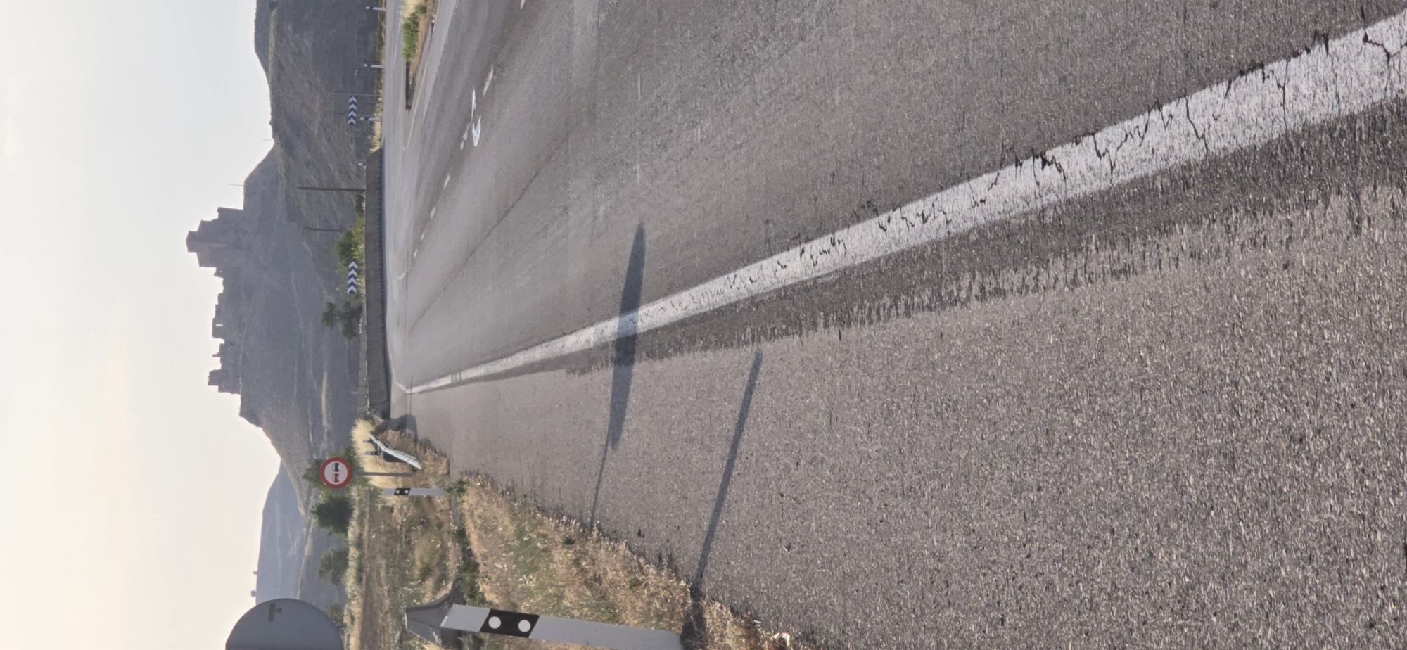 The media file shows a road in a mountainous landscape, likely in Loporzano, Spain. The road has a white line down the center and appears to be somewhat worn, with some cracking. There are roadside signs and what looks like a fortification or ruins on the hillside in the distance. The sky is bright and hazy, suggesting it is either early morning or late afternoon. The light casts a long shadow on the road, indicating the sun is low in the sky. The overall scene depicts a rural and somewhat remote location. There is a red and white circular sign with a diagonal line through it, indicating a prohibition, though the specific symbol within is unclear. Another sign with white and blue chevrons indicates a curve in the road. There are no people or animals visible in the frame. The weather appears to be clear but possibly dusty or hazy.