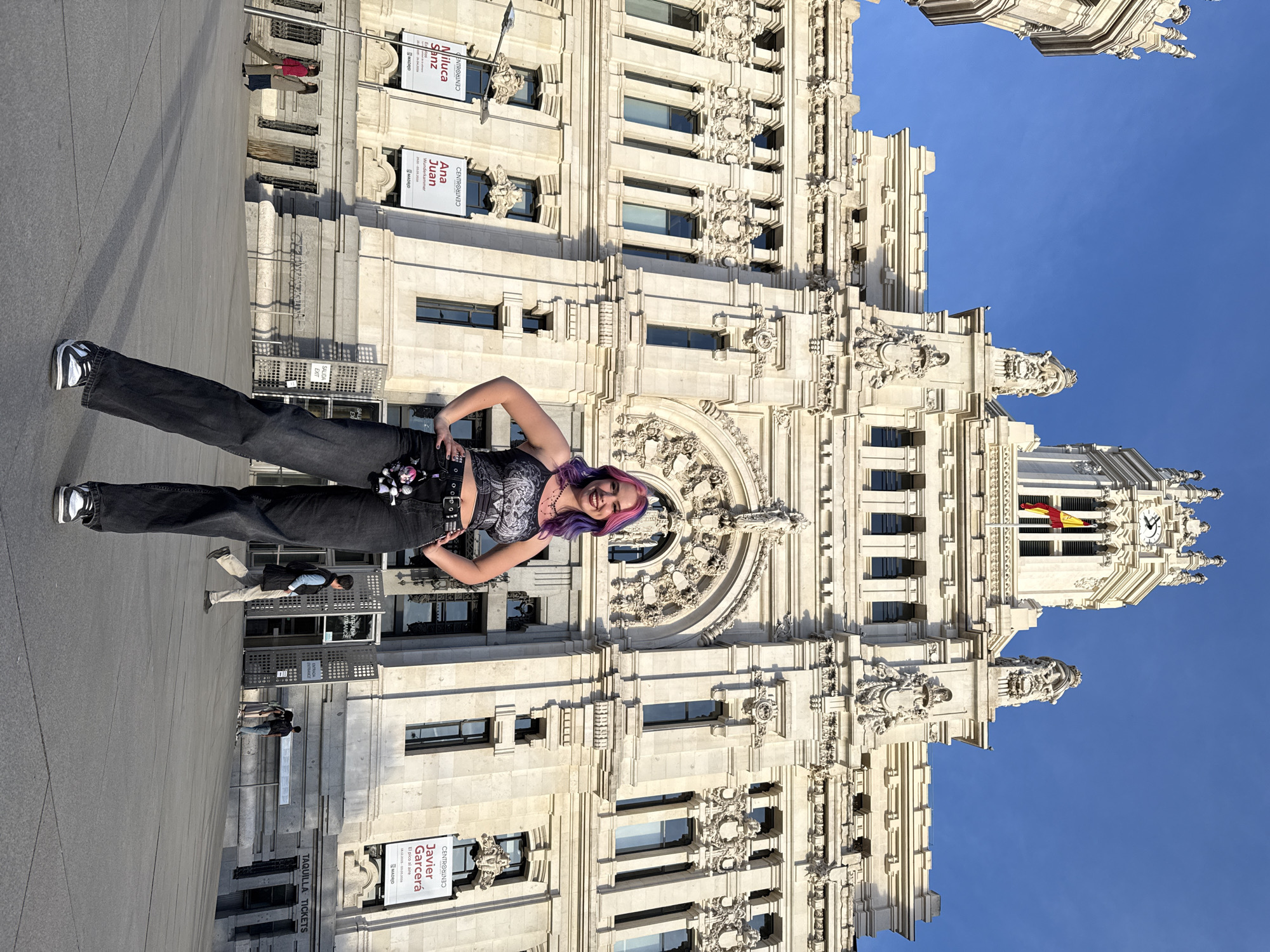 The image is of a smiling woman with purple hair, wearing black pants and a black and gray top, standing in front of a large, ornate, light-colored building. The building has many windows and decorative elements, including sculptures and carvings. Banners or posters are attached to the building. A Spanish flag is visible on top of the building. The sky is blue. People are seen walking in the background. The building is located in Madrid, Spain, as indicated by the signs that read "MADRID." The signs appear to advertise exhibitions ("Miluca Sanz", "Ana Juan", "Javier Garcerá"). There is a ticketing area called "TAQUILLA TICKETS."