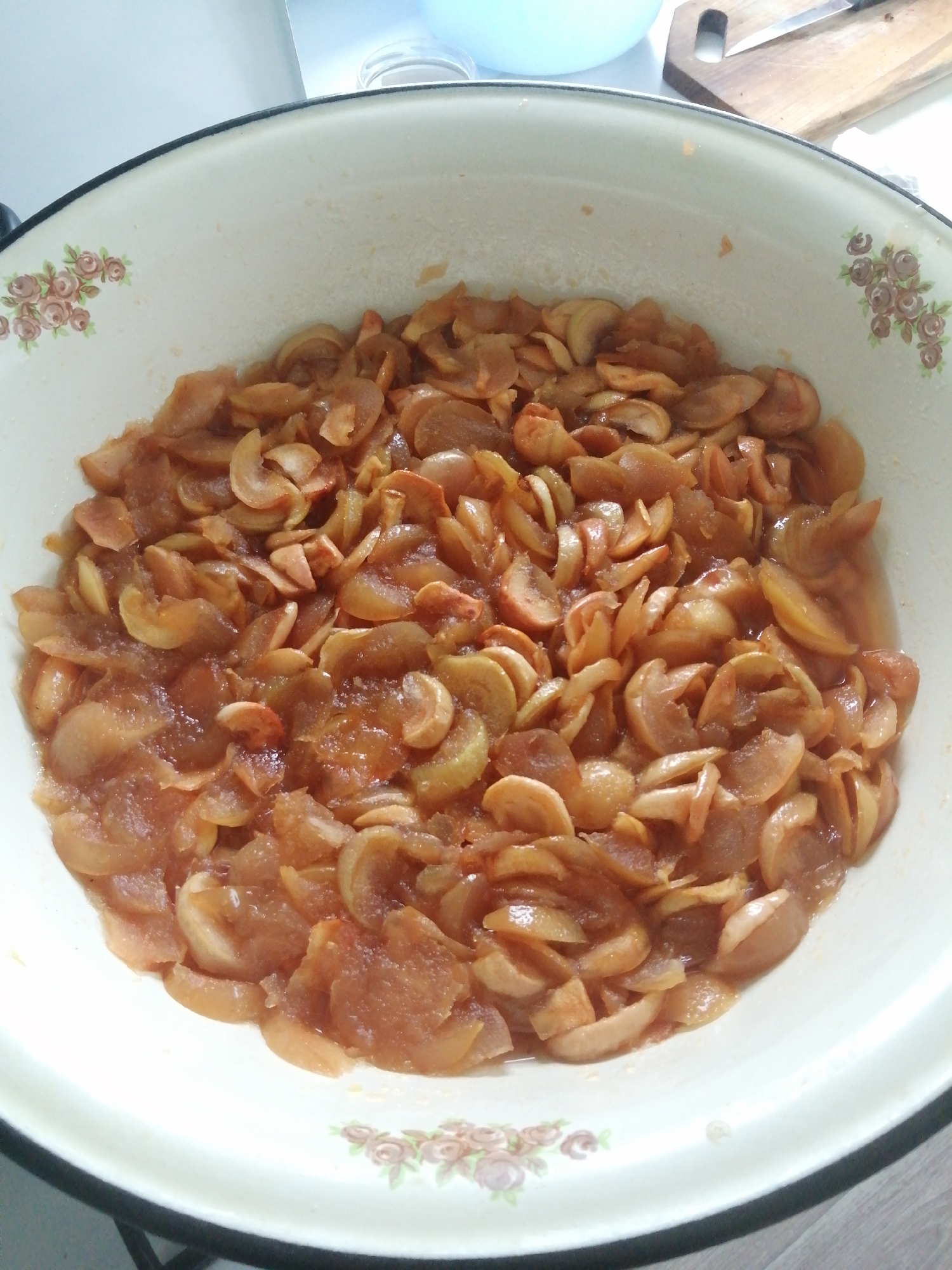 A close-up, overhead shot shows a large, shallow, enamel bowl filled with cooked apple slices. The bowl is white with a black rim and decorated with a floral pattern on the left and right sides. The apple slices are a reddish-brown color and appear soft and slightly caramelized, mixed with a syrupy liquid. The background is blurred, but suggests an indoor setting with a countertop and a wooden cutting board with a knife visible. The scene is likely from a kitchen in Slonim, Belarus.