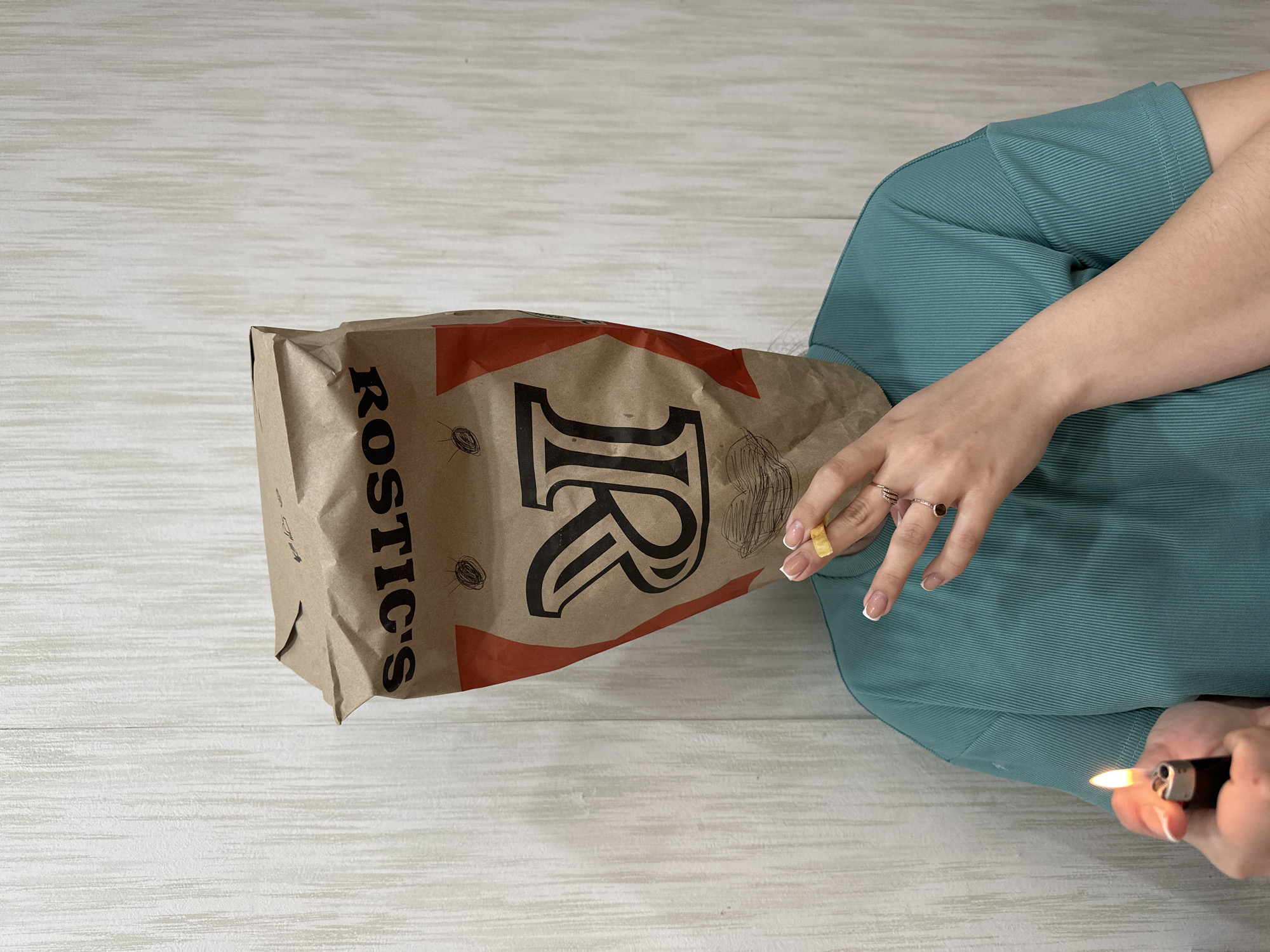 A person sitting indoors on a light-colored, wood-grain surface is holding a lit lighter in their right hand near a brown paper bag labeled "ROSTIC'S" with a large "R" on it. The bag appears to be upright. The person is wearing a ribbed, teal-colored top. A partially eaten french fry is visible in the left hand. The image was taken in Chaikovsky, Russia.
