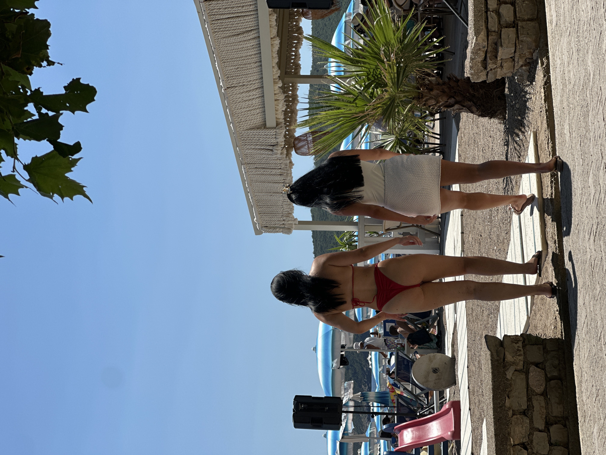 Two women are walking away from the camera on a paved walkway at a beach resort. The woman on the left is wearing a red bikini and the woman on the right is wearing a white tank top and a white skirt. In the background, there are beach chairs, umbrellas, palm trees, and a building with a thatched roof. The sky is clear and blue. The location is the beach in Boreti, Montenegro.