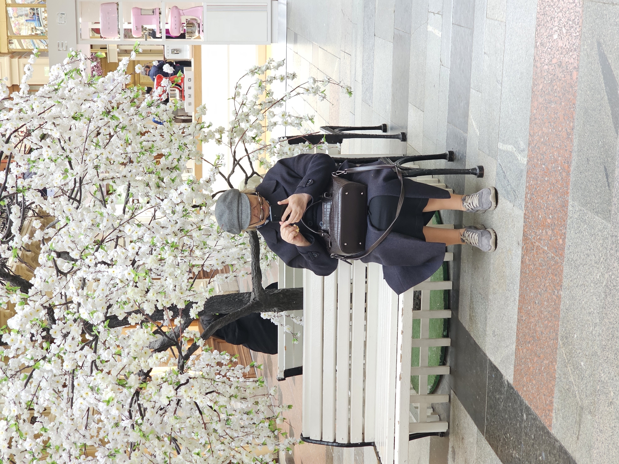 An individual in a hat, glasses, and dark clothing is sitting on a white bench, looking at a smartphone in their hands. They have a satchel resting next to them on the bench. A large artificial tree with white blossoms is beside the bench. The bench is on a tiled surface, with a store or display visible in the background. The photo appears to be taken indoors in an urban environment.