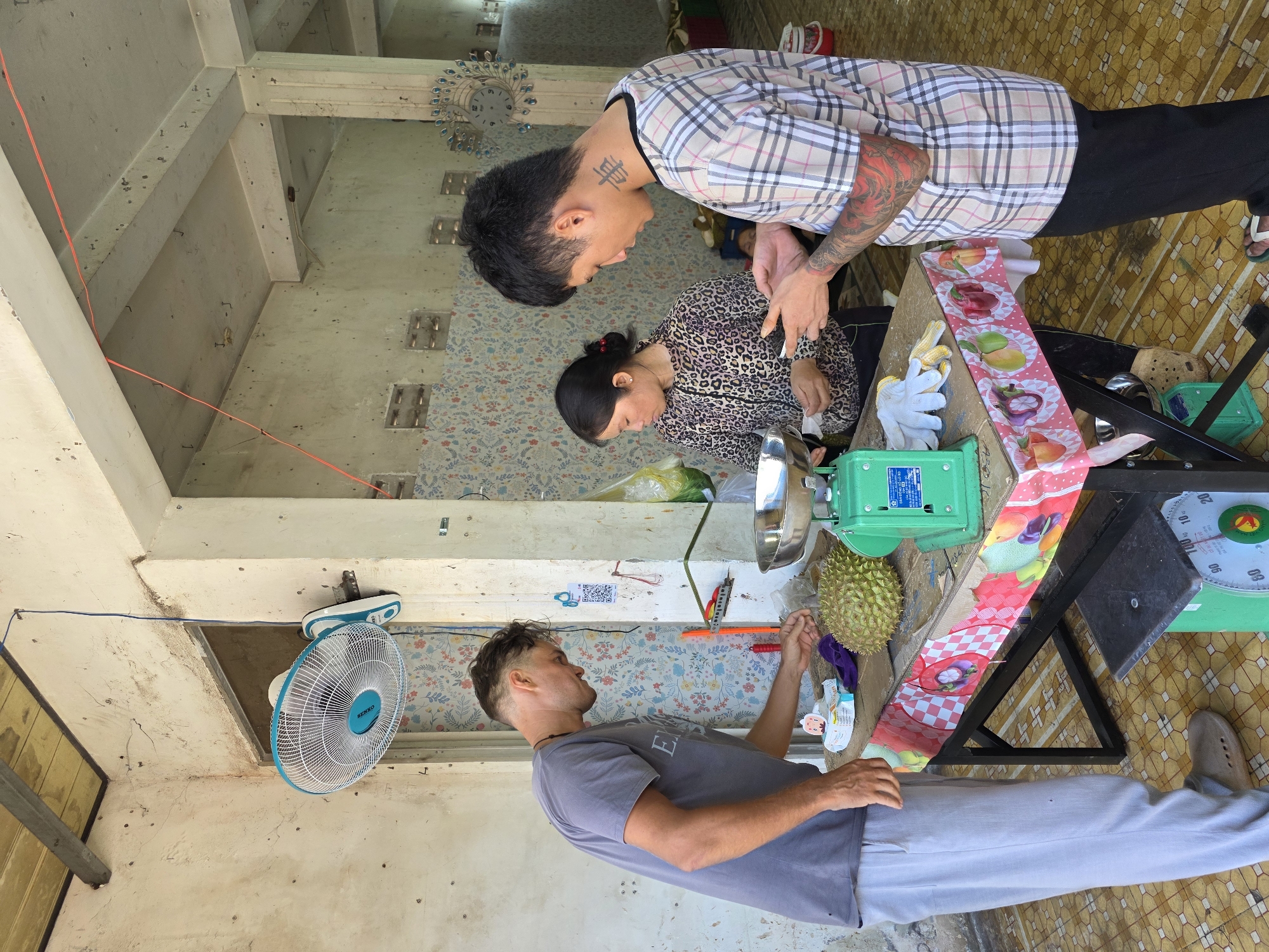 The image captures an outdoor scene in Phan Thiết, Vietnam, likely a market or roadside stall, with a focus on three individuals and several objects. Two men and one woman are present. The woman, wearing a leopard-print top, stands behind a table. A man with tattoos on his arm and neck, wearing a plaid shirt, leans in towards her, seemingly engaged in conversation. Another man, wearing a gray t-shirt and light-colored pants, stands facing the table, his gaze directed towards a durian fruit.
On the table, a green electronic scale and a circular analog scale are visible, suggesting the weighing of produce. A large durian fruit is placed near the scales. A decorative strip with images of various fruits like apples, mangoes, and peppers is draped over the table. A white glove lies on the table, possibly for handling the fruit. A blue and white oscillating fan is mounted on the wall in the background. The setting appears to be an open-air structure with concrete beams and a patterned tiled floor. The lighting suggests it is daytime. The overall mood seems casual, with the individuals interacting in a routine manner.