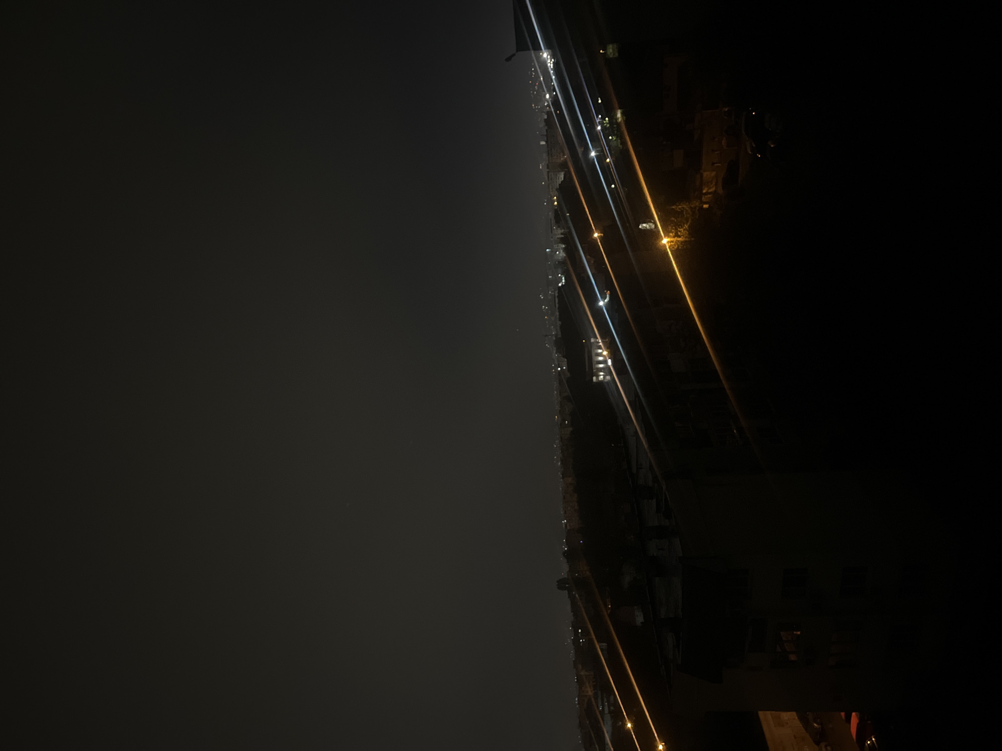 The image captures a nocturnal panoramic view of a city, identified as Sofia, Bulgaria, from an elevated perspective. The scene is characterized by deep darkness punctuated by artificial illumination. The upper portion of the frame is dominated by a dark, monochrome sky, likely overcast or heavily influenced by light pollution, as no celestial bodies are visible.
A long, horizontal band of scattered lights marks the distant cityscape, indicating the extent of urban development stretching into the horizon. These pinpricks of light reveal the presence of buildings, streets, and infrastructure.
The immediate foreground, particularly towards the lower right, consists of dark, indistinct silhouettes of structures, possibly parts of buildings or rooftops close to the camera.
Cutting diagonally across the image are several vivid streaks of light, predominantly bright white and warm yellow/orange. These appear to be lens flares or internal reflections caused by intense, nearby artificial light sources, rather than physical elements within the distant urban fabric. They create a dynamic visual element against the otherwise static backdrop.
The overall atmosphere is one of quiet vastness, showcasing the spread of a city after dusk. No specific activities, individuals, or identifiable landmarks are apparent. There is no visible text.