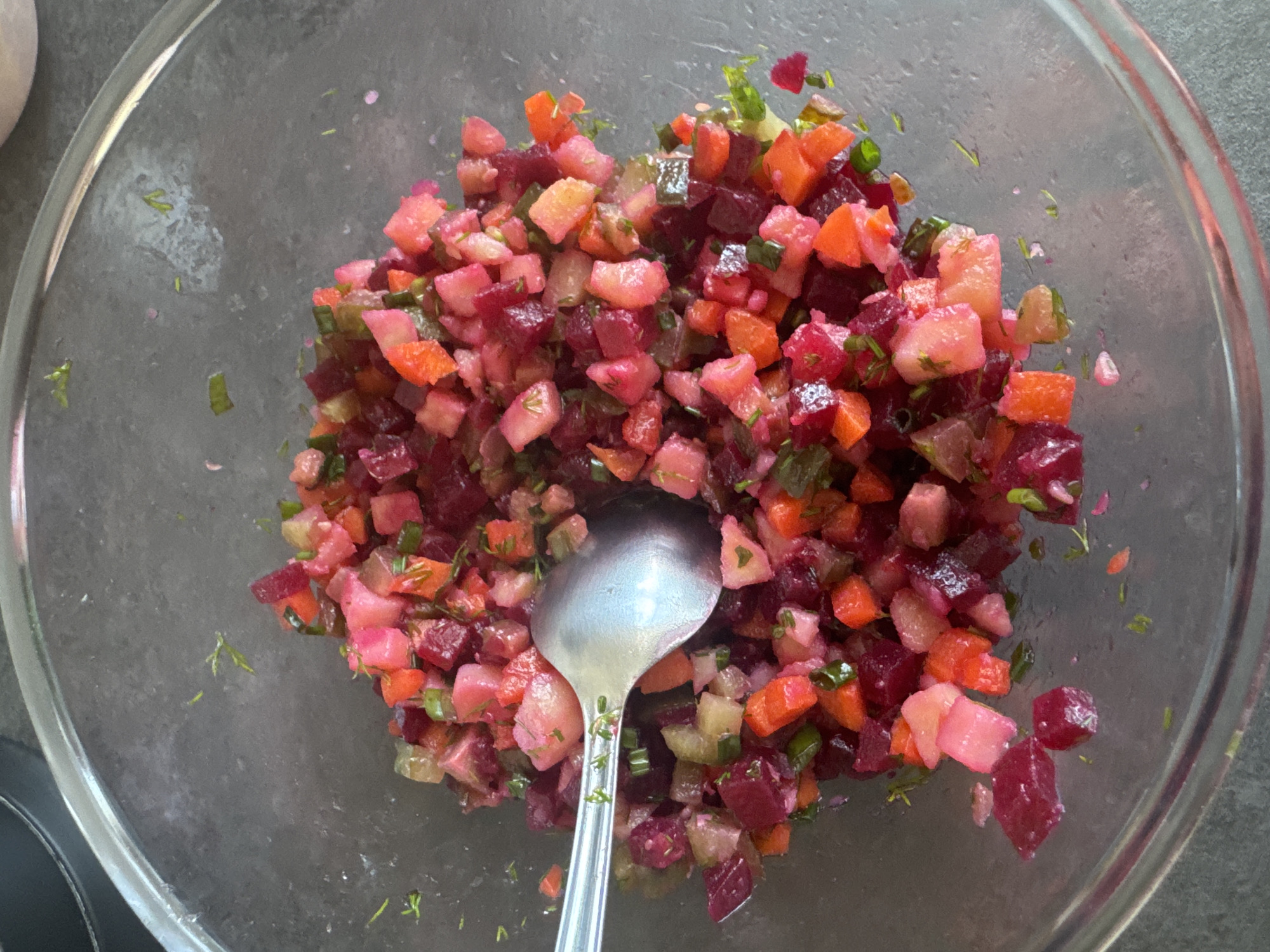 The scene, located in the city of Chaikovsky, Russia, presents an overhead view of a clear glass bowl filled with a diced vegetable salad. The salad comprises approximately 1-2 cm cubed pieces of dark red beets, light pinkish-beige potatoes, orange carrots, light green pickles, and fine green herbs. A silver-colored metal spoon is partially submerged within the salad, positioned diagonally from the upper left. The glass bowl is situated on a dark grey surface, with a blurred dark object visible in the upper left background.