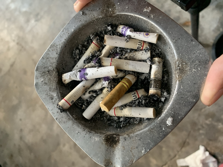 The image captures an overhead view of a well-used metallic ashtray filled with numerous cigarette butts and scattered grey ash. A person's hands are visible on the top-left and right edges, holding or presenting the ashtray.
The ashtray is square-shaped with rounded corners and indentations for resting cigarettes, showing signs of frequent use with accumulated residue. The cigarette butts are predominantly white-filtered, with various markings. Many feature purple bands with visible English text such as "CLICK" and "MN," along with Thai script (e.g., "บุหรี่" meaning "cigarette"). One white-filtered butt has a red band with a stylized "B" visible. A distinct golden-brown cigarette butt, differing in color from the others, lies among the white ones. Another butt features a black patterned filter. The ends of the cigarettes are dark and burnt, indicating they have been smoked.
The background is a blurry, light grey surface, consistent with a concrete floor, with a dark, indistinct object partially visible on the right. The lighting is uniform, suggesting an indoor or sheltered daytime setting. The scene depicts the aftermath of smoking, with the ashtray serving as a receptacle for the finished cigarettes. The presence of Thai script on the cigarette butts aligns with the provided context of the location being Metro, Indonesia, indicating popular brands in the region.