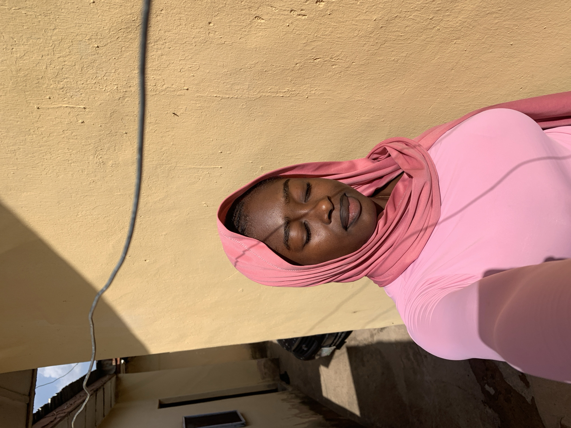 A person wearing a pink top and head covering lies supine against a beige-painted wall. A power cable is visible along the wall, above a balcony. The surroundings are likely residential buildings. The location is Rafin Par, Nigeria.