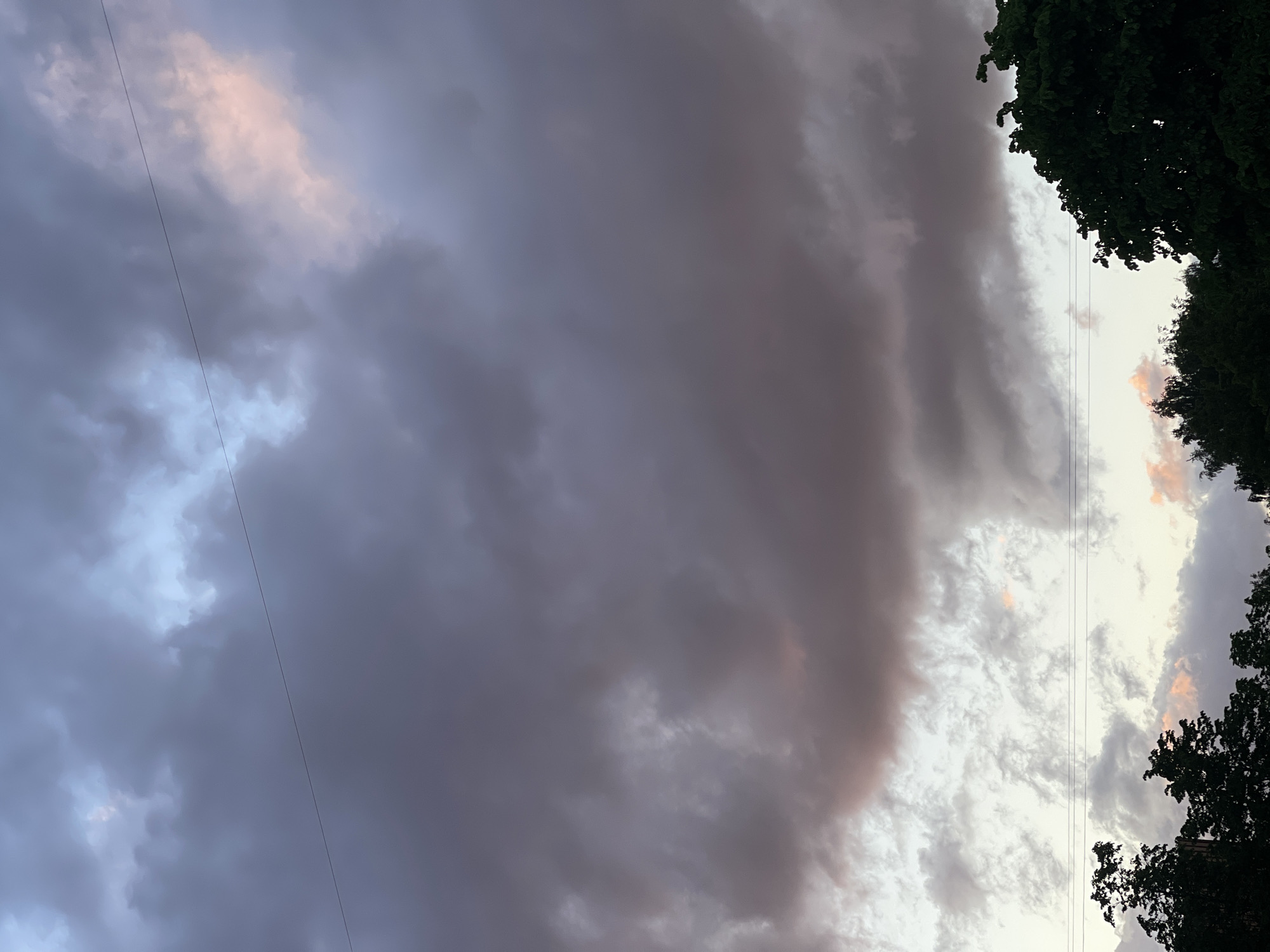 The image shows a sky filled with storm clouds, with tree foliage visible on the right side. Electric cables run vertically through the scene. There are traces of pink light in the clouds. Without further context, the location in Riga, Latvia, cannot be identified forensically, as there are no indicators (landmarks, signage, architectural styles) that would tie this scene to a specific location within the city.
