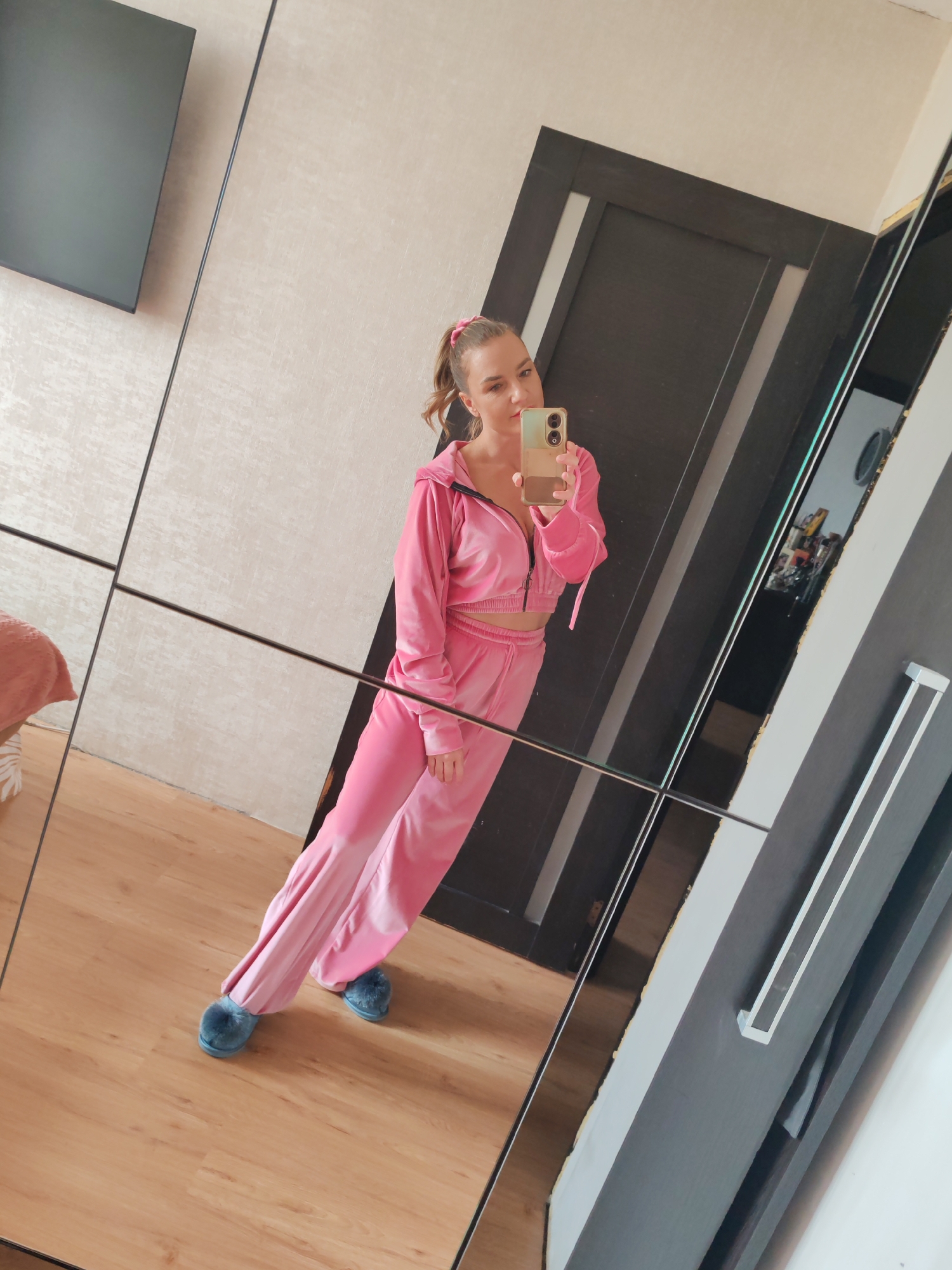 A woman is standing in front of a mirrored wardrobe, taking a selfie with a phone. She is wearing a pink velour tracksuit and blue fuzzy slippers. Her hair is pulled back in a ponytail with a pink scrunchie. The wardrobe has a black door with a white trim. The floor is wooden. A grey television screen can be seen on the wall. Due to the reflection and lack of landmarks, the location in Petropavlovsk-Kamchatsky, Russia, cannot be determined.