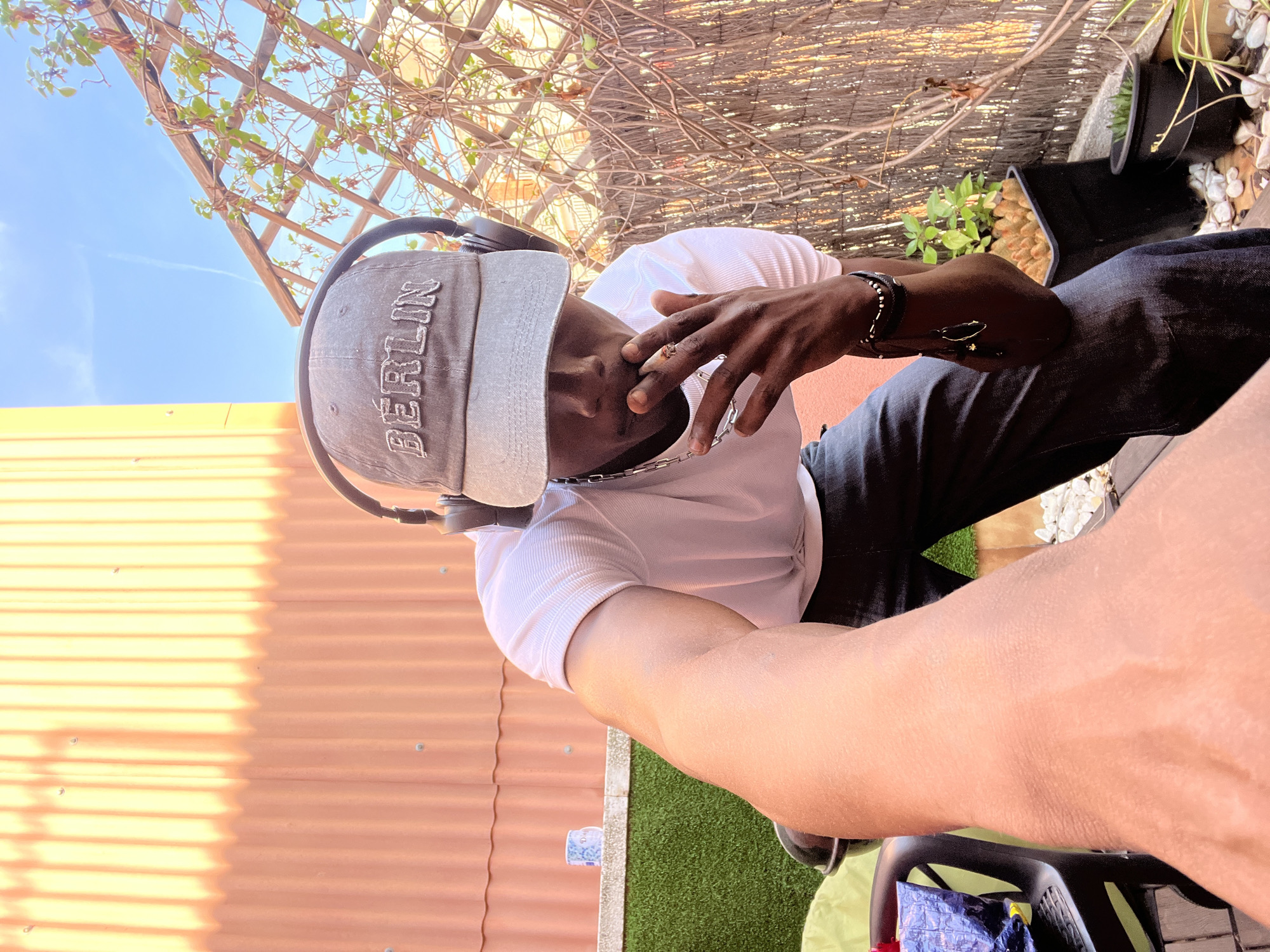 The image presents a man outdoors, possibly in a garden or patio setting. The location is possibly in the city of Barcelona, Spain as the cap he is wearing has "Berlin" stitched on it. The man is wearing a white t-shirt, dark jeans, and a hat with the word "BERLIN" embroidered on the front. He also has headphones around his neck and is wearing jewelry on his wrist and neck. His hand is raised near his face in a thoughtful gesture. The background includes a fence, plants, and a glimpse of the sky. A chair and a green surface are visible in the foreground.