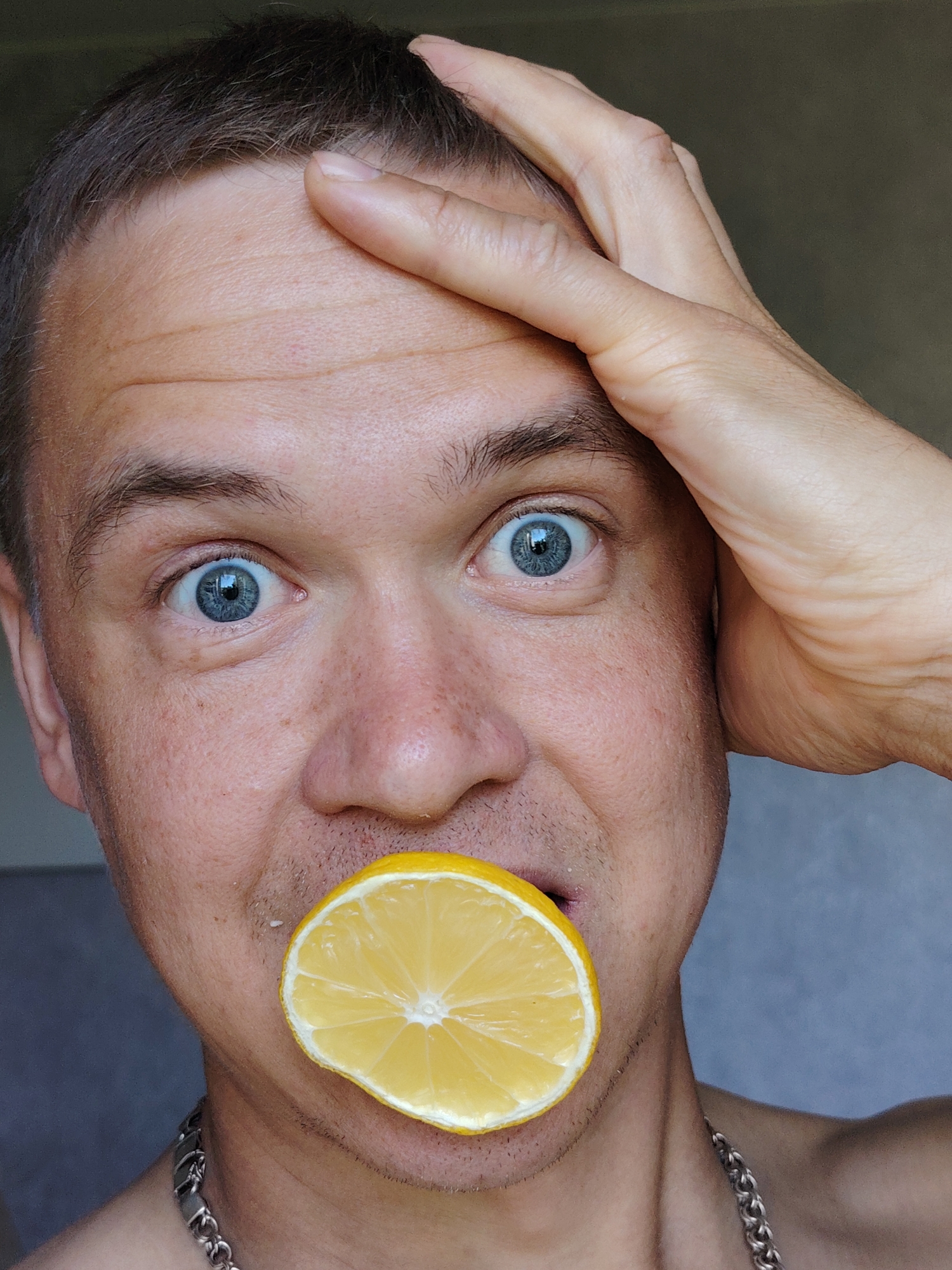 A close-up photograph shows a Caucasian male with short brown hair, light skin, and blue eyes holding a slice of lemon in his mouth. His right hand is resting on his forehead. He appears to be indoors, judging by the neutral-colored wall in the background. A silver chain is visible around his neck. The details don't provide enough information to confirm the location is Chaikovsky, Russia.