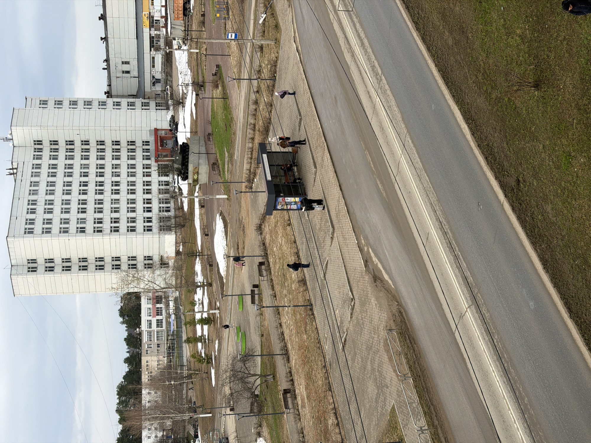 The image shows an urban scene in Chaikovsky, Russia, from an elevated perspective. A large, white apartment building dominates the left side of the frame. Adjacent to the building is a pedestrian walkway and a road. People are visible on the walkway and near a bus stop or transit shelter with advertisements. The road appears to have tram tracks running along it. In the distance, more buildings and green spaces can be seen. The season appears to be early spring, with patches of snow still present on the ground.