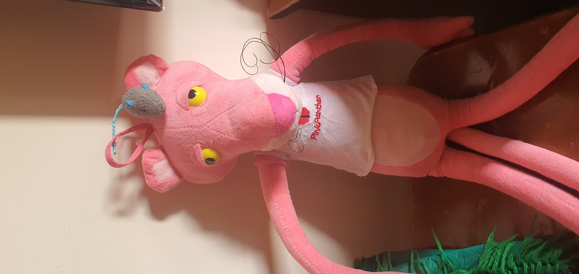 A stuffed pink panther toy is sitting on a brown surface, likely a table or shelf. The toy has yellow eyes and is wearing a white t-shirt with "Pink Panther" embroidered on it in red. A small grey mouse toy is attached to the top of its head with a blue string and a pink loop. The background is a plain wall with shadows cast from the toy and other unseen objects. To the left of the toy, a corner of a green fuzzy object, possibly a scarf or blanket, is visible. The scene is indoors, and the lighting suggests it is daytime, with light coming from the right. No specific location in Baku, Azerbaijan can be determined from the image.