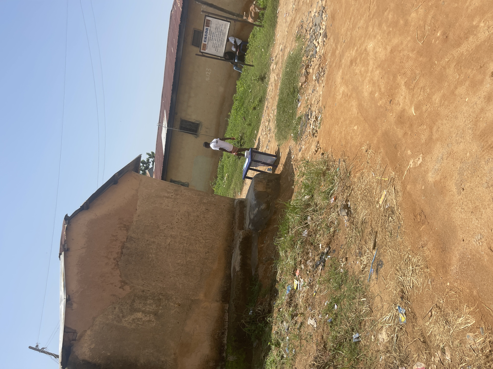 The image depicts an outdoor scene, likely in Jos, Nigeria. A light-colored, stucco-walled building with a corrugated metal roof dominates the left side of the frame. In front of this building, a person in a white t-shirt and red shorts stands near a blue and white plastic object that appears to be a table or a water dispenser.
Further back, a larger building with a similar roof and wall construction has a sign posted on it. This sign has text, but it is mostly illegible in the image. Some of the visible text includes "CASOR" and "N.123". Two other individuals, also appearing to be wearing white tops, are seated near this sign. A goat or similar animal is also visible behind them.
The ground in front of the buildings is a mix of dirt, sparse grass, and some debris, including pieces of plastic and paper, suggesting it might be a somewhat unkempt area. The sky is clear and blue, indicating a sunny day. The orientation of the image is tilted, making it appear as if the scene is on its side. The overall atmosphere is casual and appears to be daytime.