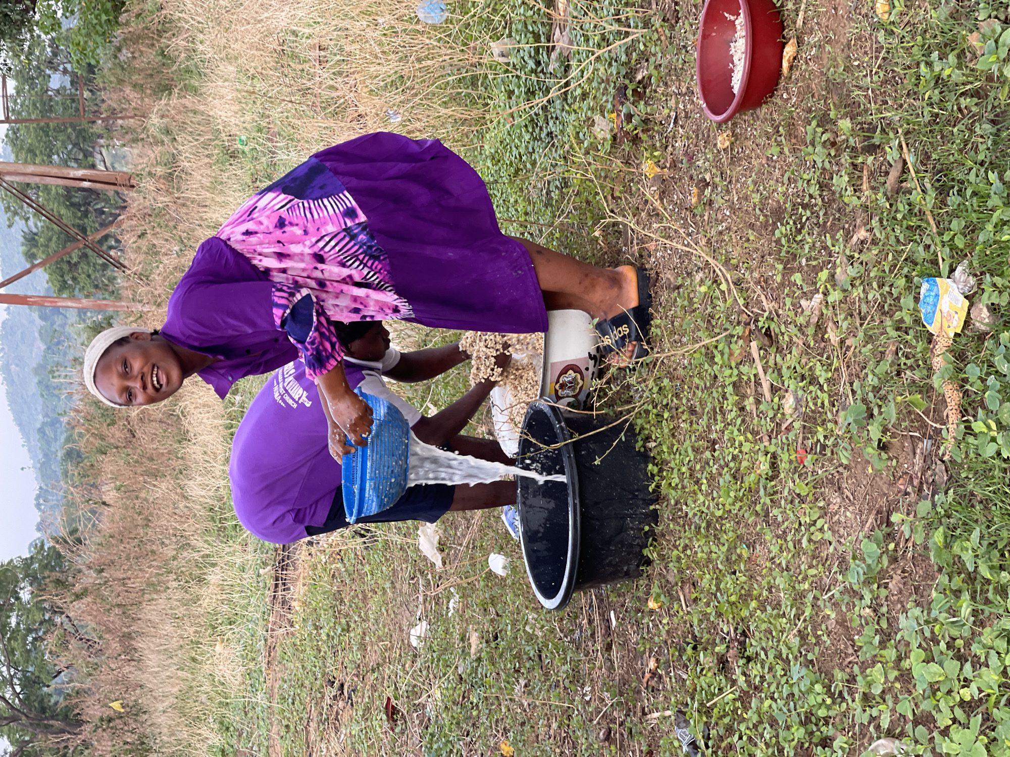In an outdoor setting in Tabu, Nigeria, a woman is bent over pouring water from a blue plastic colander into a black plastic bucket. The woman is wearing a purple dress with a pink and white patterned top, a white head covering, and black Adidas slides. She is smiling and looking at the camera. Next to her is a white container filled with what appear to be nuts or seeds. A second person is partially visible behind the woman. A red bowl containing white matter is in the upper right corner. The ground is covered with a mix of brown grass, green foliage, and scattered trash. A hill with trees and power lines is visible in the background.