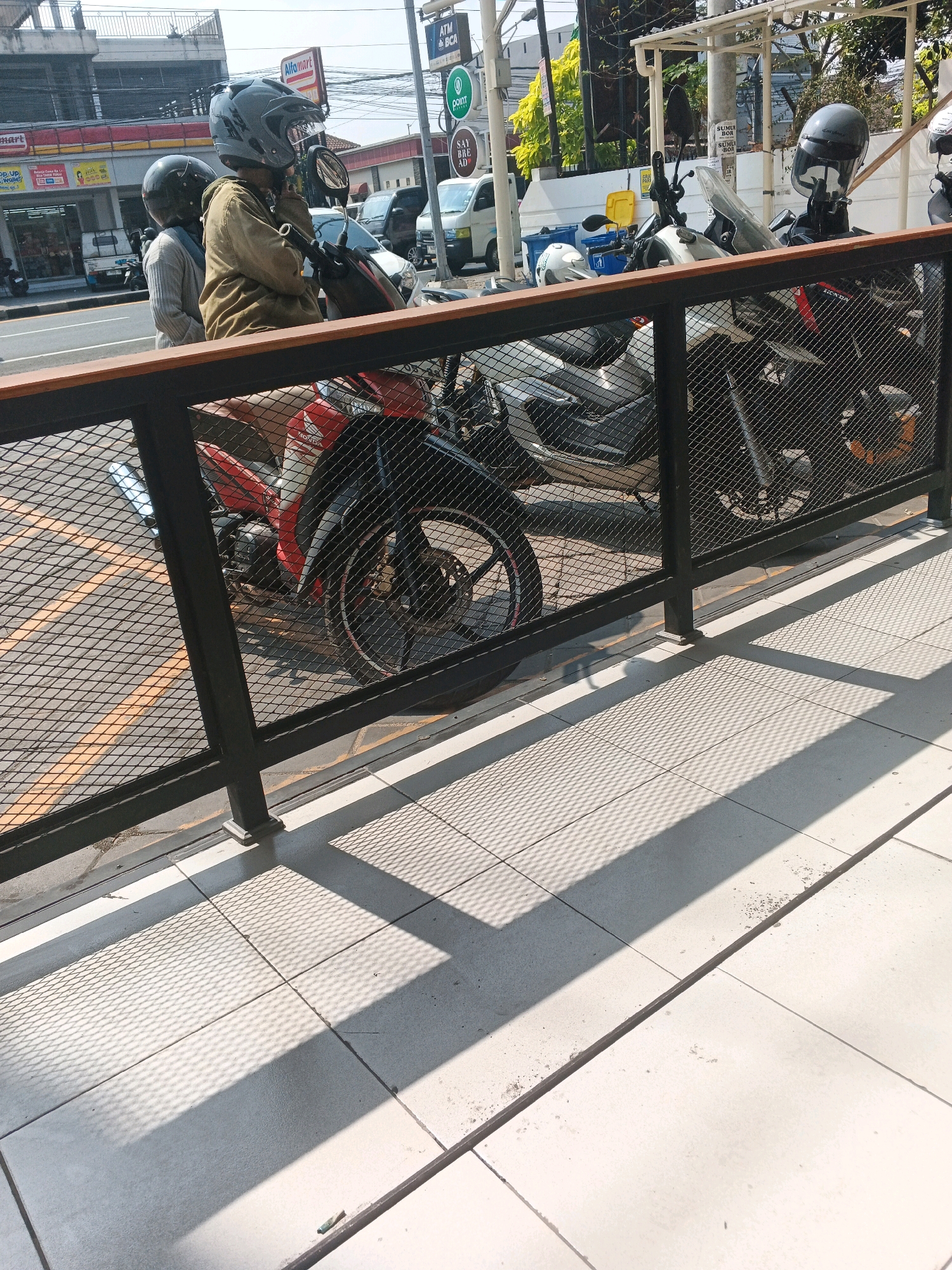 The image depicts an outdoor scene in Genuk Barat, Indonesia, likely a street or sidewalk area adjacent to commercial establishments. In the foreground, a black metal fence with a diamond mesh pattern runs horizontally across the frame. Sunlight casts a distinct shadow pattern from the fence onto the light-colored tiled ground.
Behind the fence, several motorcycles are parked. One motorcycle, predominantly red and black, is partially visible, with its front wheel and fender in focus. To its right, another motorcycle, black with a windshield, is parked. Further back, another motorcycle, primarily black, is visible.
In the background, two individuals are seated on a motorcycle. The person in front, wearing a dark grey helmet with a face shield, is dressed in a brown jacket. The person behind them is wearing a black helmet. They appear to be either waiting or preparing to depart. Beyond the parked motorcycles and people, a white van and a white car are visible, along with a red and white building that appears to be a store with signage. Traffic lights and street signs are also present in the background, indicating a public road. The overall environment suggests a typical urban or suburban setting in Indonesia.