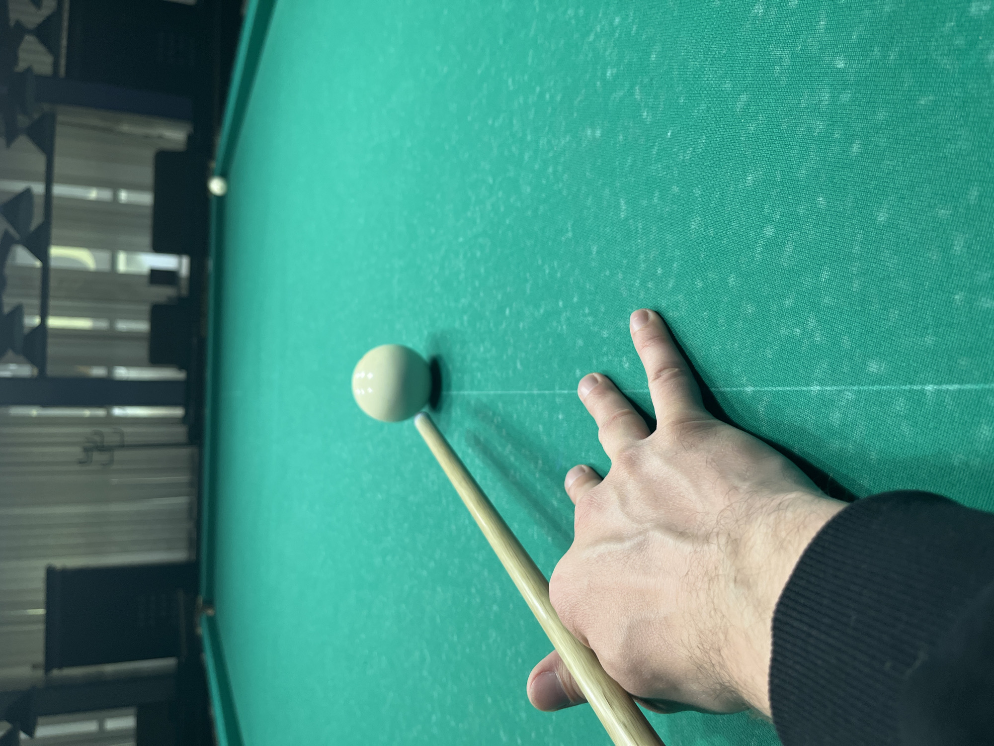 A person is playing pool on a green felt pool table. A cue stick points towards a white cue ball. The person's hand rests on the table, close to the cue ball. The setting appears to be indoors, possibly a recreation room or pool hall. The location is Unknown, Unknown.