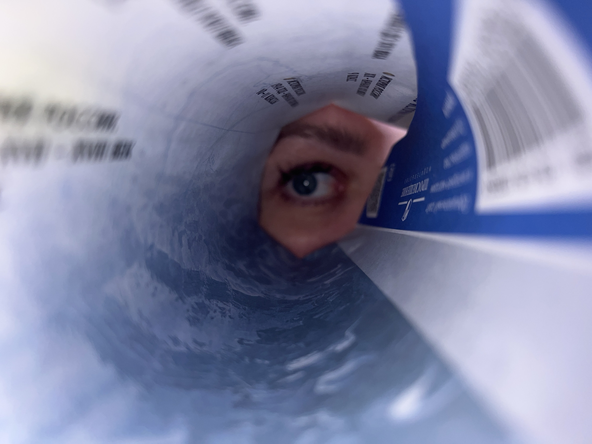 A subjective, extreme close-up view from within a curved, cylindrical object, likely a rolled document or container. A human eye with a prominent blue iris, partial eyelid, and eyebrow is visible in the mid-ground, peering directly forward. The interior surface of the object is light-toned, featuring blurred black Cyrillic text. Recognizable text fragments include "ИСТОРИЯ РОССИИ" (History of Russia), "9 КЛАСС" (9th Grade), "1914 ГОДА" (1914 Year), and "10-11 КЛАССЫ" (10-11 Grades), indicating the content is related to Russian educational materials. To the right, a segment of the object's exterior or an adjacent panel is visible in a solid blue color, displaying blurred vertical lines consistent with a barcode. The bottom interior of the object exhibits a dark blue and grey swirling texture, resembling ripples or a stylized pattern. The scene is evenly illuminated. No specific geographical landmarks or features within the city of Noviy, Russia are discernible from the image to pinpoint an exact location beyond the presence of Russian-language educational material.