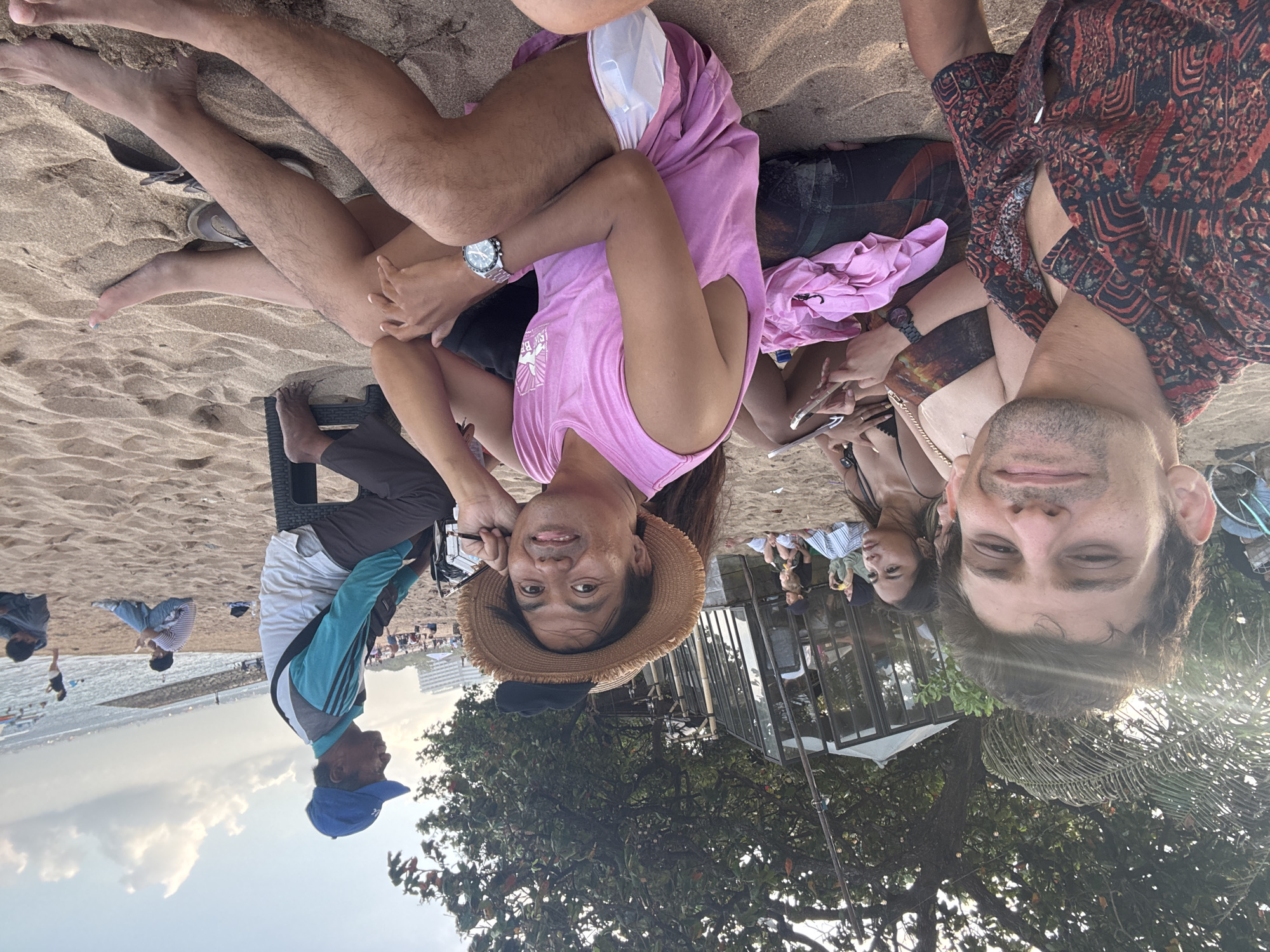 In Denpasar, Indonesia, on a sandy beach, a group of people are gathered. In the foreground, two men and two women are seated on the sand. One man in a patterned shirt is in the left foreground. Beside him, a woman wears a bikini top. Another man in a pink tank top and a straw hat is in the center, facing the camera. A woman in a patterned dress sits to his right. In the background, a vendor in a blue cap and turquoise and white shirt sits on a black stool. Behind them, several other people are visible on the beach and in the ocean. A glass-enclosed structure with trees and foliage behind it is also present. The sky is clear with scattered clouds, and the sun is setting, casting a warm glow on the scene.