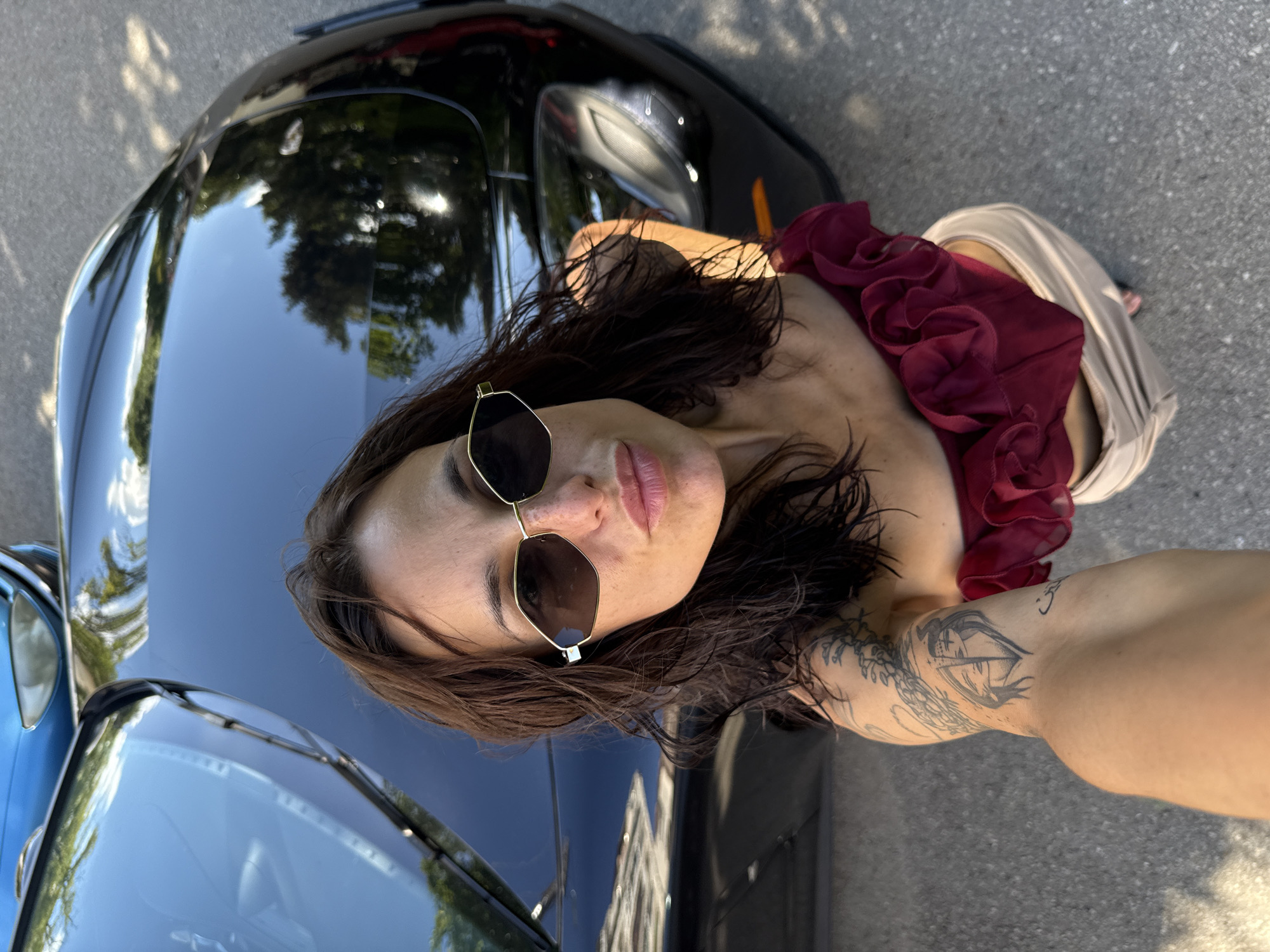An overhead, full-body selfie shows a woman standing next to a black car on a paved surface. The woman has dark, wavy hair and is wearing sunglasses, a dark red ruffled top, and light-colored pants. A tattoo is visible on her left arm. The car appears to be parked on a street, with trees and other cars visible in the reflection of the black car's hood and roof. The lighting suggests it is daytime. The image's metadata does not contain location information, therefore, the city of Pleven, Bulgaria, is not ascertainable from the image.
