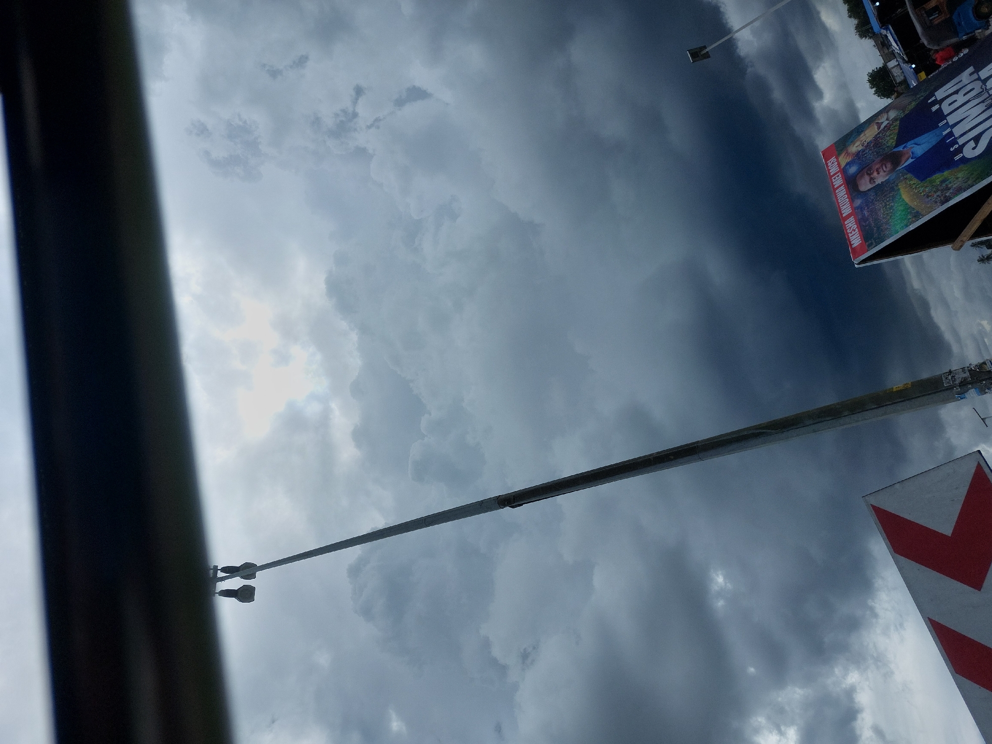 The image presents a view of a cloudy sky in Dar es-Salaam, Tanzania. A streetlight extends towards the center of the frame. A billboard with the name "SIMBA" is visible on the right side of the image. The lower right corner displays a white and red chevron-shaped road sign. A blurred dark object is present on the left side of the image. The sky is dominated by dark, heavy clouds.