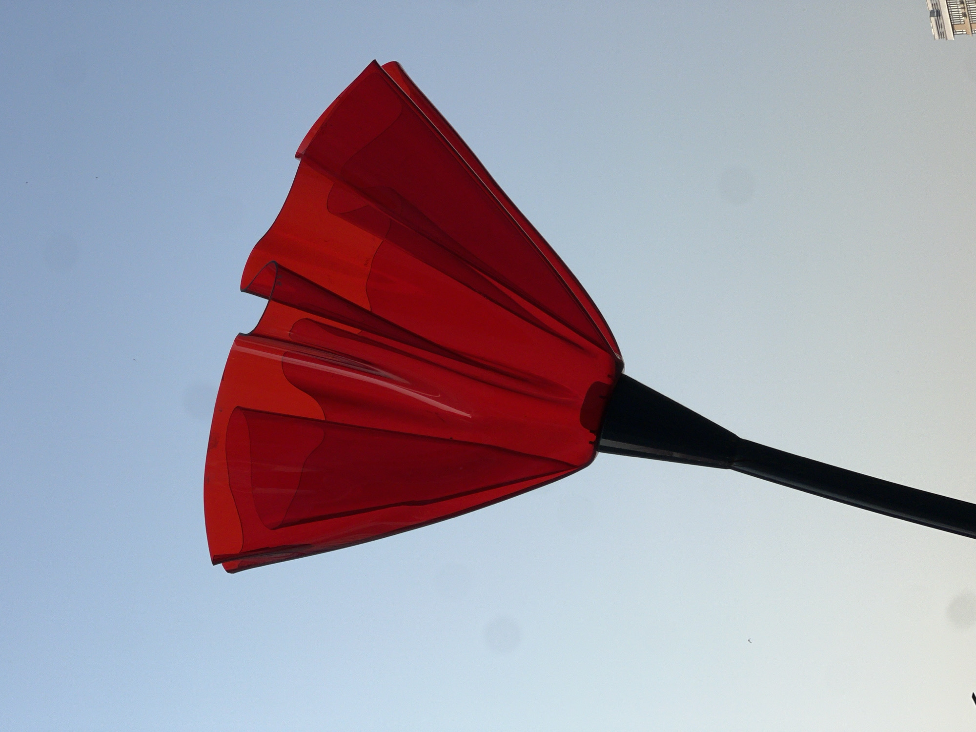 A red, translucent umbrella is angled against a clear sky, with its fabric spread open. The umbrella is held by a black pole. In the distance, a building with horizontal white bands is visible at the bottom right of the frame. The scene is likely located in the city of Krasnodar, Russia.