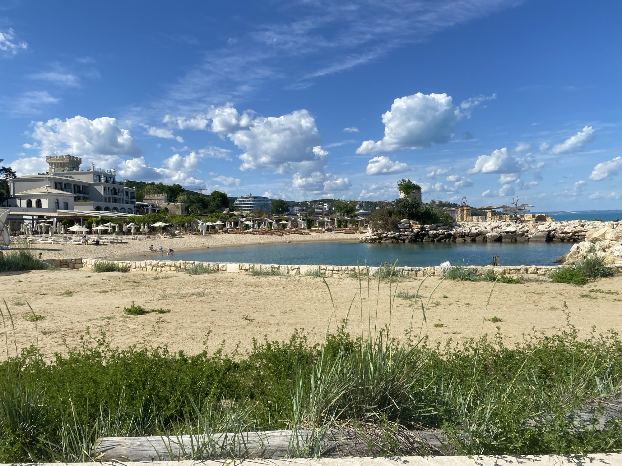 This photograph captures a sun-drenched beach scene in Varna, Bulgaria. The foreground is dominated by coarse sand and interspersed with patches of low-lying green vegetation and tall grasses, partially obscured by a weathered wooden beam. Beyond this, a sandy expanse leads to a small, enclosed bay with calm, azure water, lined with stone embankments. In the background, a resort area is visible with numerous white umbrellas shading beach chairs, suggesting a relaxing vacation spot. Architectural structures, including a building resembling a castle tower, add character to the landscape. In the distance, hints of urban buildings are nestled among lush trees. The sky is a vibrant blue, filled with scattered, puffy white clouds. Several people are on the beach, engaged in various activities such as sunbathing and playing near the water's edge.