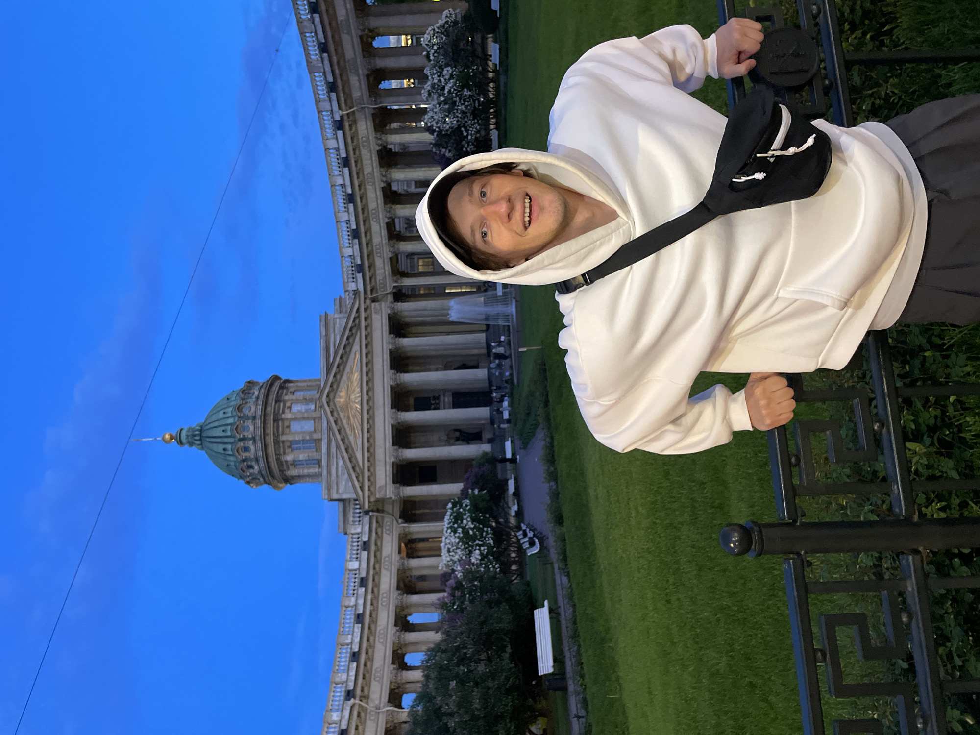 A young man is captured in a vibrant outdoor portrait, smiling broadly at the camera during twilight hours. He is positioned on the right side of the frame, wearing a white hooded sweatshirt and a black cross-body bag with white drawstrings. He holds onto a decorative black metal fence with both hands, which runs horizontally across the foreground.
In the background, a grand neoclassical building with a distinctive green dome, reminiscent of the Kazan Cathedral, rises majestically. Numerous tall columns line its facade, and a fountain can be seen in the middle distance in front of the building. Lush green grass stretches across the scene, interspersed with trees, some bearing white blossoms. The sky is a clear, deep blue, indicative of dusk or early evening, and some artificial lights illuminate the cathedral. A thin wire or cable is visible stretching across the sky. The man's cheerful expression suggests a happy and relaxed mood. While there appears to be a raised emblem on the fence post, any text is indistinct. The setting is consistent with Saint Petersburg, Russia.