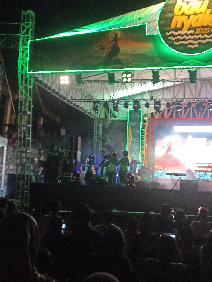 The image captures an outdoor night event, likely a concert or cultural performance, in Kecamatan Pujut, Indonesia. A large, complex stage structure, constructed from metal trussing, dominates the scene, bathed in bright green illumination.
On the stage, several performers are visible, some dressed in green attire, amidst stage lights and a light haze or smoke effect. The performers appear engaged in an act, suggesting an ongoing show. Above the main stage, a prominent rectangular LED screen displays a striking graphic: a silhouetted figure, possibly a dancer or mythical creature, against a vibrant, fiery background. To the upper right, a large circular banner or sign features the text "bau nyale 2025" with wavy lines underneath, indicative of a festival or event name scheduled for the year 2025. Another bright LED screen is positioned on the right side of the stage, also displaying visuals.
The foreground is filled with the dark silhouettes of a large audience, gathered to watch the performance. Their heads and shoulders are visible, indicating a significant crowd. Some audience members appear to be holding up phones, potentially recording the event or taking photos. The dark sky above confirms the nighttime setting, with all illumination originating from the stage lighting and screens. The overall impression is that of a lively public gathering for a cultural or entertainment event.