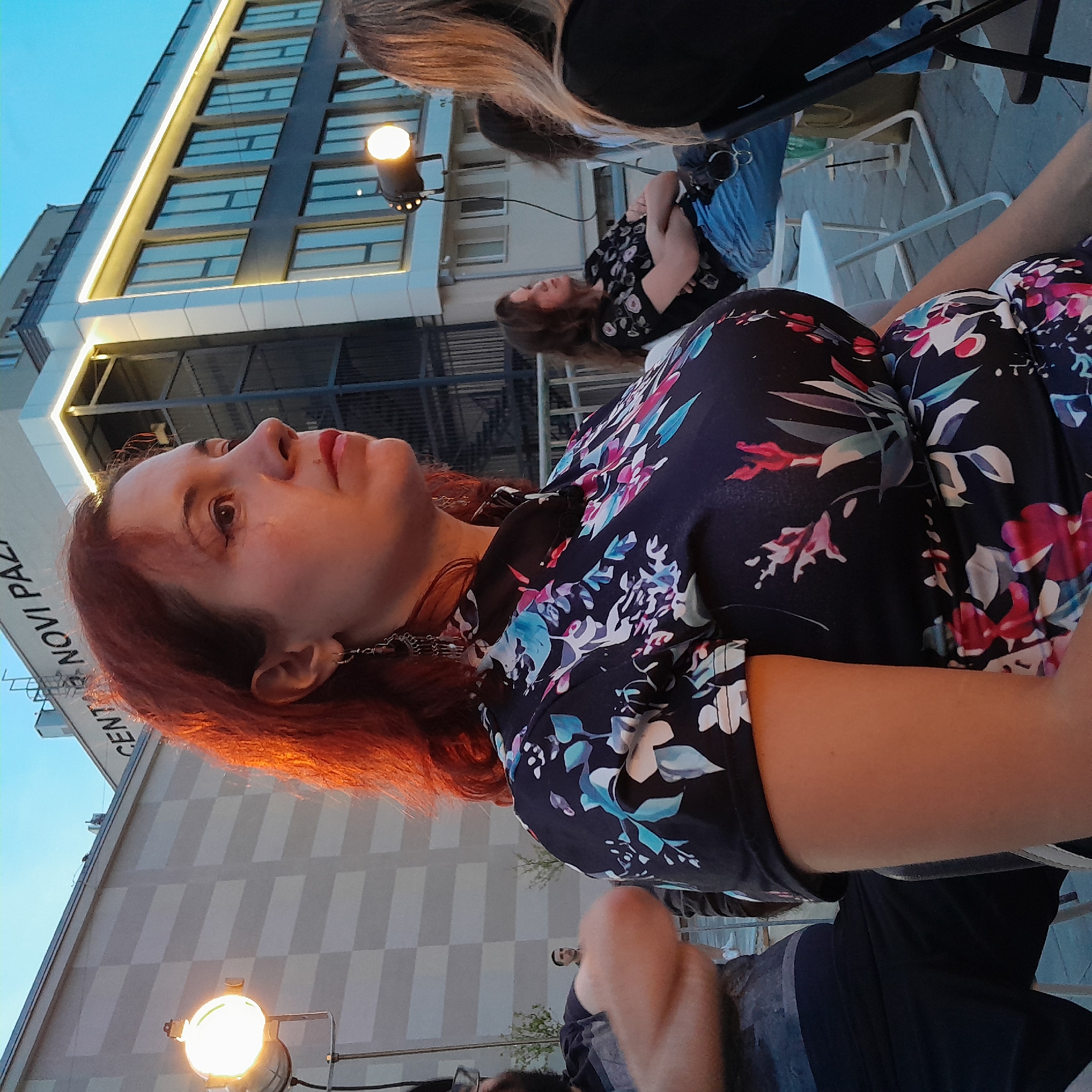A woman with auburn hair and a floral dress is seated outdoors, facing right, with a building in the background. The building has a checkered pattern on its facade and the words "CENTAR NOVI PAZAR" are visible at the top. There are also lights illuminating the building and spotlights on the ground. Other people are visible in the background, some seated and some standing. The lighting suggests it is late afternoon or early evening.