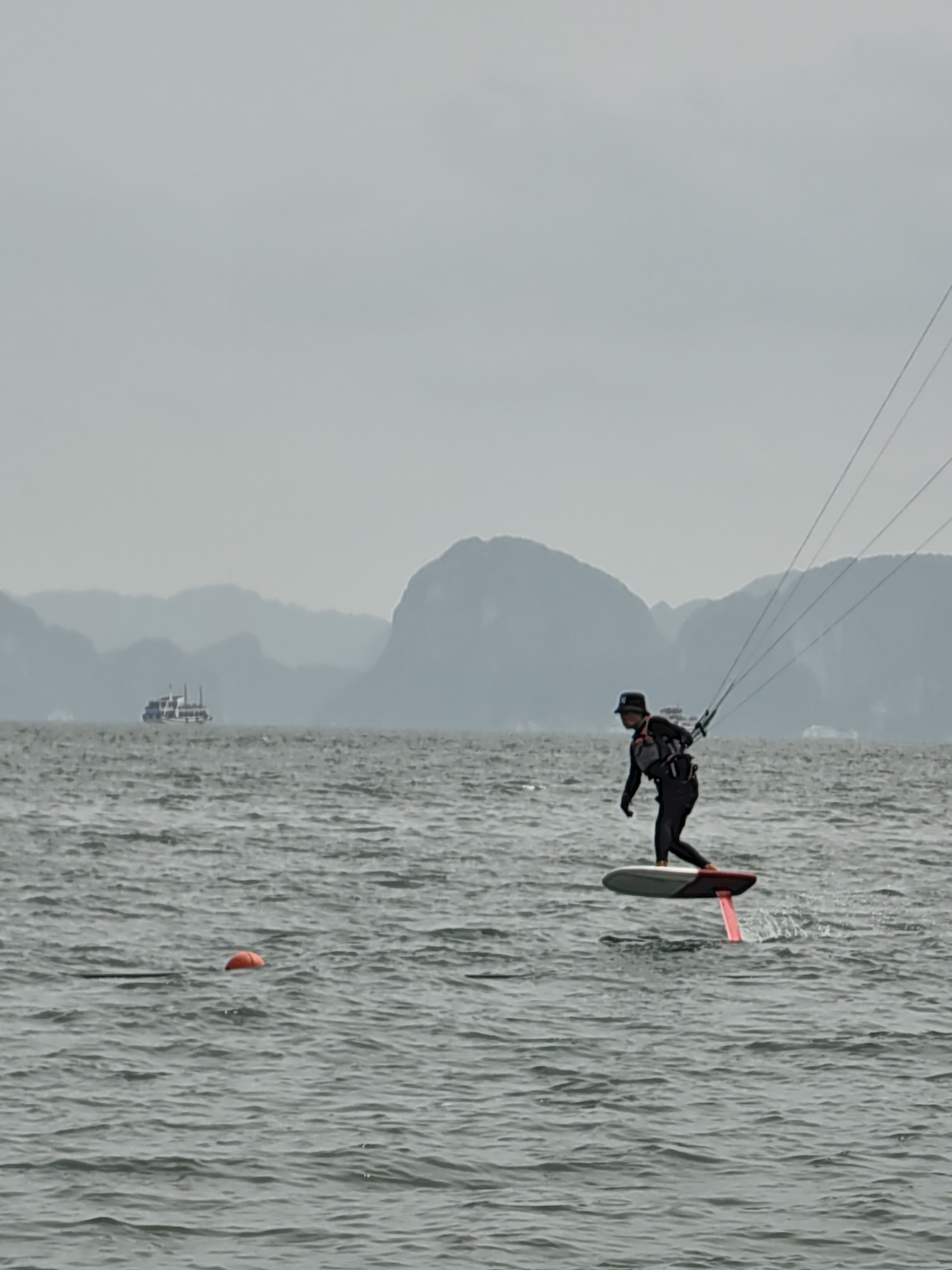 A person is kitesurfing on the water in Bai Chay Ward, Vietnam. The individual is wearing a black wetsuit, life vest, and a hat. They are standing on a foilboard, with a red hydrofoil wing visible beneath the board. The kite lines are extending upwards and out of frame. In the background, there are hazy mountains and a boat. An orange buoy is floating in the water to the left. The water has a slight chop to it.