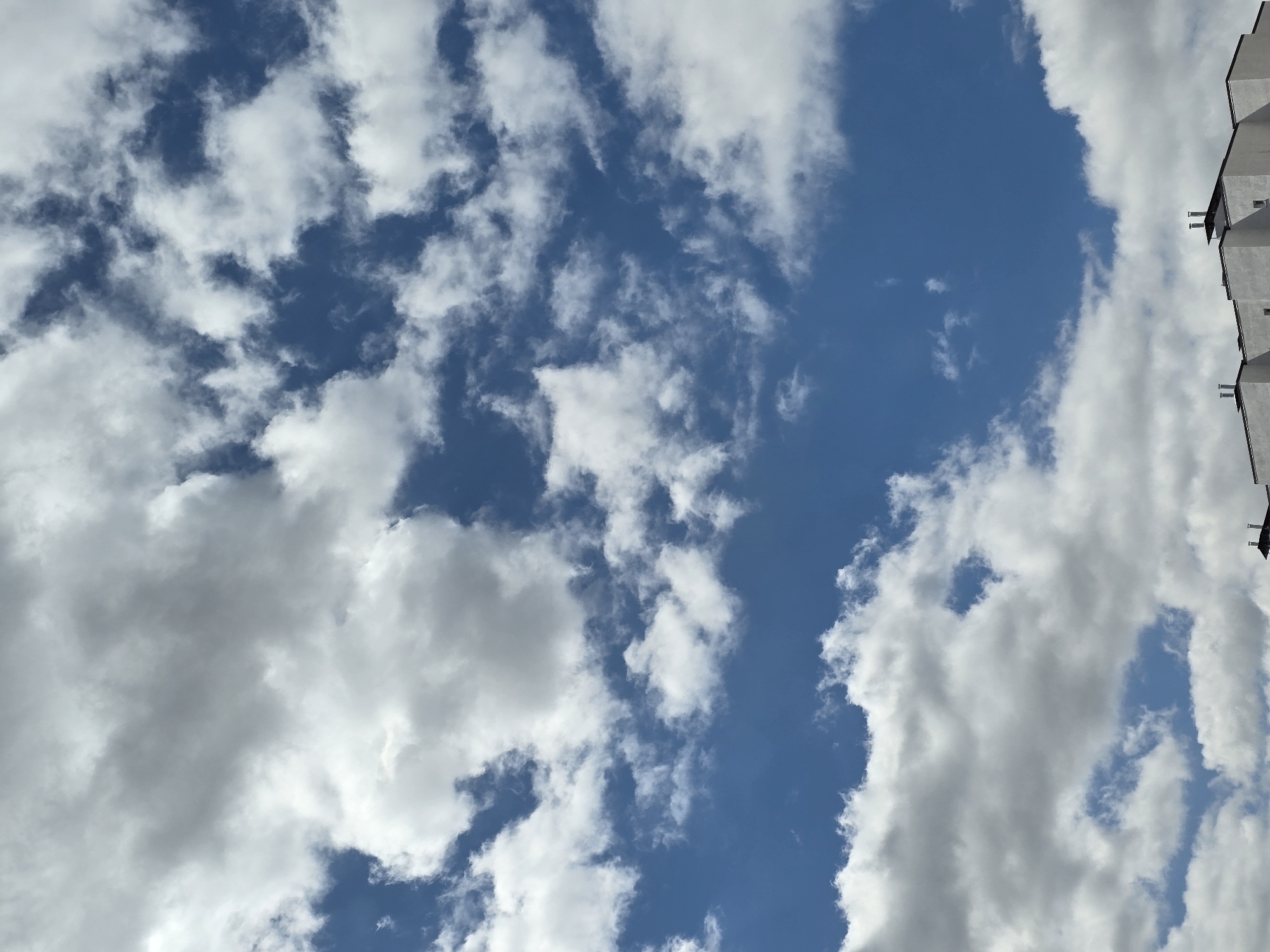 The image shows a clear blue sky with scattered white cumulus clouds, taken from a low angle looking upwards. The bottom portion of the image features the roofline of a modern, white, multi-story building with several chimneys. The building appears to be located in a developed urban or suburban area. The sky is the dominant element, suggesting an outdoor daytime scene, possibly in Sofiivska Borshchahivka, Ukraine. There are no people or animals visible in the frame, and no actions are taking place. The environment is characterized by the contrast between the bright sky and the architectural structure below.