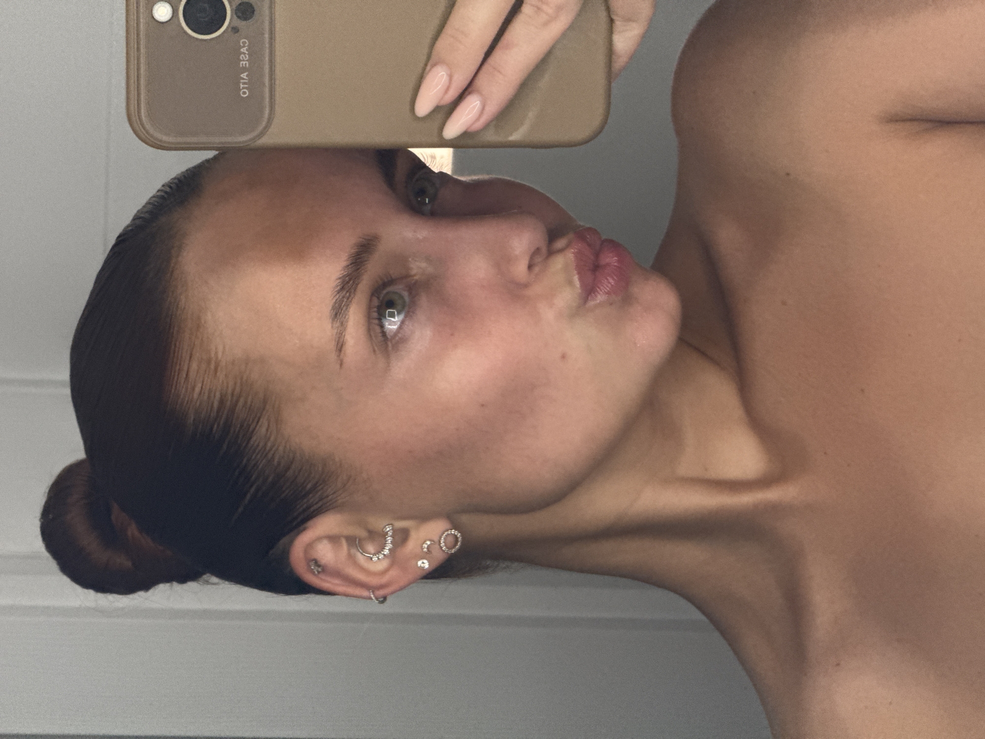 This media file captures a close-up mirror selfie of a young woman, presented rotated 90 degrees clockwise. The woman, shown from the chest up, has fair skin, dark hair pulled back into a neat bun, and light-colored eyes. She is making a playful pouting or "kissing" expression towards the camera.
Her left hand, adorned with long, light pink, almond-shaped artificial nails, holds a light brown or beige smartphone. The back of the phone and its camera module are visible at the top of the frame. Reversed text can be seen on the phone case, including "OTIA 3ZAJ" and "CAGE VLLO".
Her left ear is prominently displayed, featuring multiple piercings: a helix piercing with a curved, jeweled bar, a small hoop in the conch, a tragus stud, and various lobe piercings including studs and a circular, jeweled earring. The woman appears to be shirtless or wearing a strapless garment, highlighting her shoulders and collarbones. The background is out of focus, suggesting a simple, light-colored indoor setting.
The image depicts the activity of taking a self-portrait in a mirror. No specific time of day or weather conditions are indicated, and there are no immediate visual cues within the image itself that link to the stated location of Chaikovsky, Russia. The overall mood conveyed is one of confidence and playful self-expression.