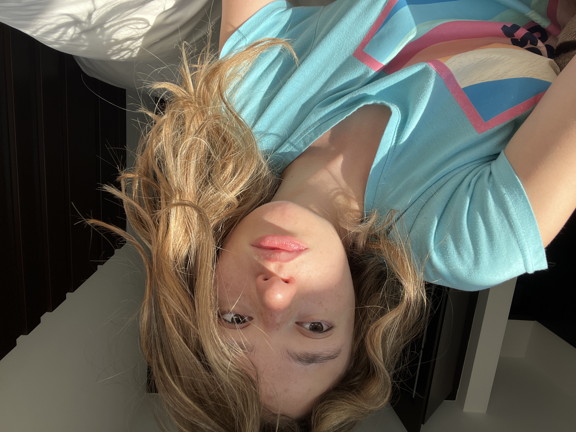 A young woman with light blonde hair is lying down, facing the camera, wearing a light blue t-shirt with a geometric design that includes pink, orange, blue and white. Her eyes are brown, and she has freckles. The background includes what appears to be white bedding, dark brown vertical panels, and white wall. The image is taken with a slight upward angle towards the subject, rotated 180 degrees, and no specific indication relates to the city of San Bernardino Tlaxcalancingo, Mexico.
