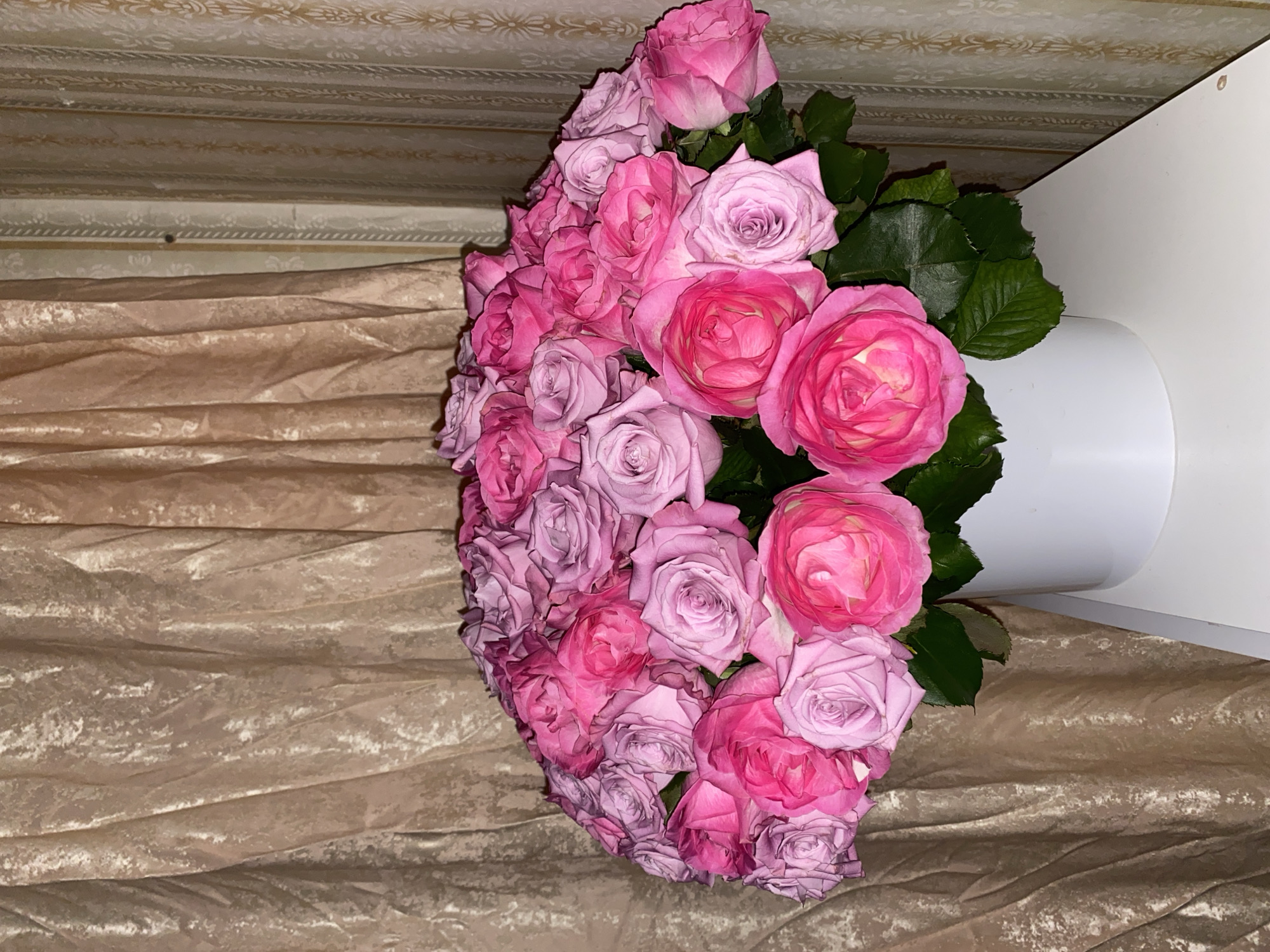 A bouquet of pink and purple roses is displayed in a white vase on a white surface. The vase is positioned in front of a beige, textured curtain. To the right of the curtain, there is a patterned wallpaper. The lighting in the image is dim. The scene is indoors and there are no people present. The image does not provide any information about the city of Lomonosov, Russia.