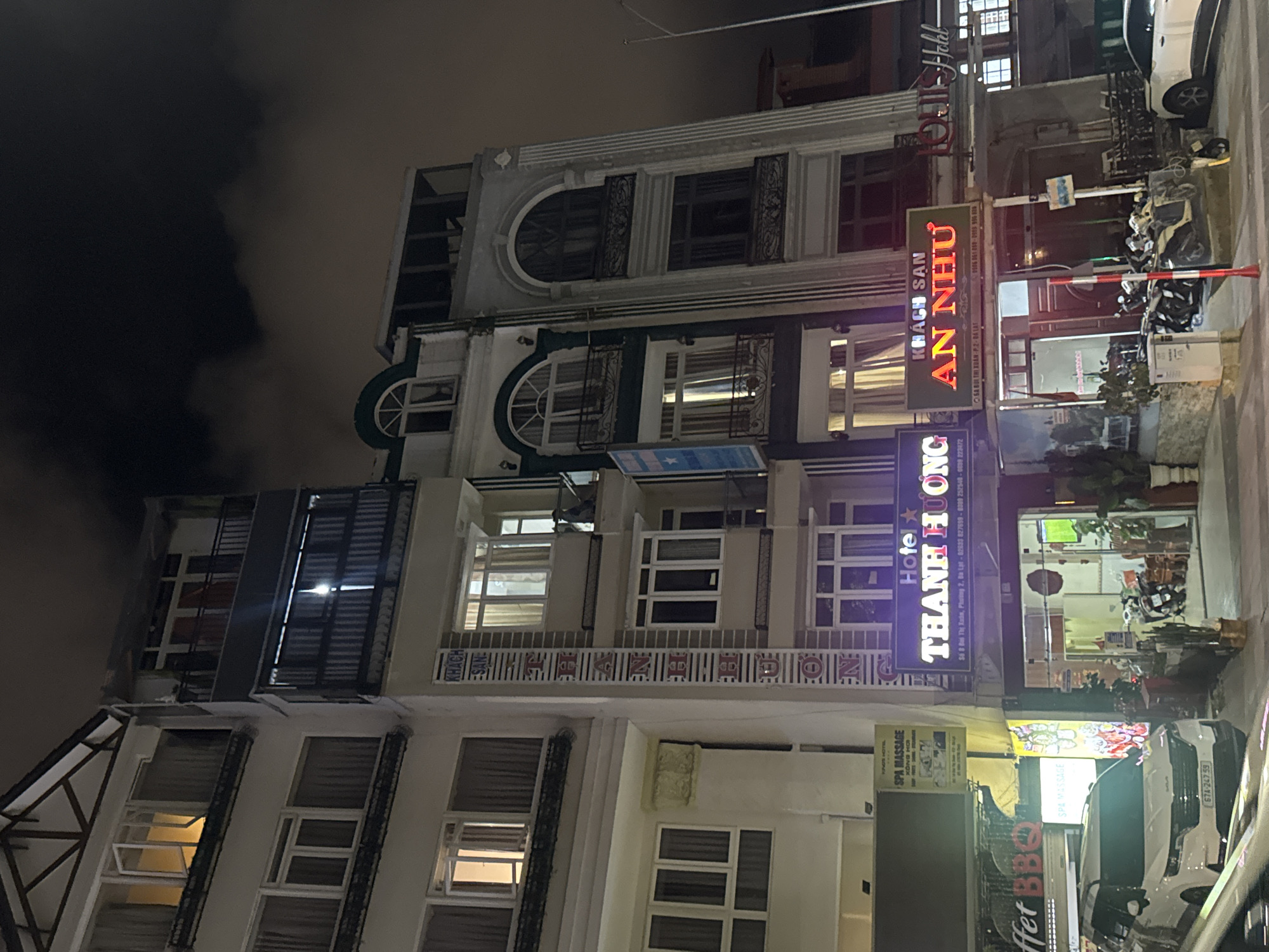 The image shows a multi-story hotel building at night in Da Lat, Vietnam. The hotel appears to be named "Thanh Huong Hotel" and "An Nhu Hotel" based on illuminated signs. The architecture is a mix of styles, with some balconies featuring ornate railings. Windows are illuminated from within, suggesting occupancy. In front of the hotel, several motorcycles are parked, and a white SUV is visible on the left. Signs for "Spa Massage" and "Buffet BBQ" are also present, indicating other businesses operating in the vicinity. The street is paved, and a street lamp is visible on the right side of the image. The sky is dark with some clouds.