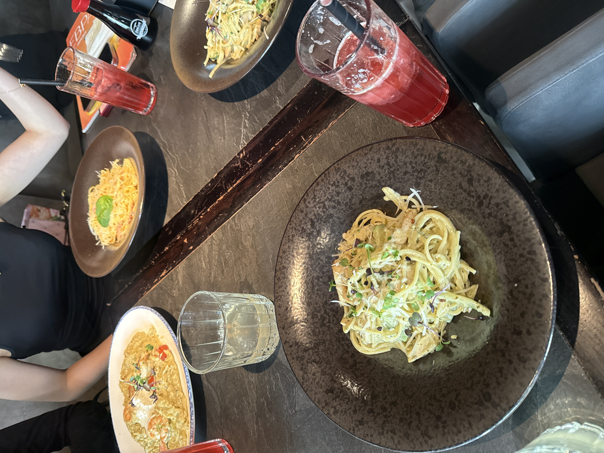The image shows a dining table with several plates of pasta dishes and drinks. Four plates of pasta are visible, each with a different style and presentation. There are two drinks, one a clear liquid in a glass and the other a red beverage with a straw in a tall glass. A bottle of soy sauce is also present. The table appears to be in a restaurant or dining establishment. Part of a person is visible on the left side of the image. This image may have been taken at the address of 194044, Russia, St. Petersburg, Bolshoy Sampsonievsky Ave., 45 литера А.