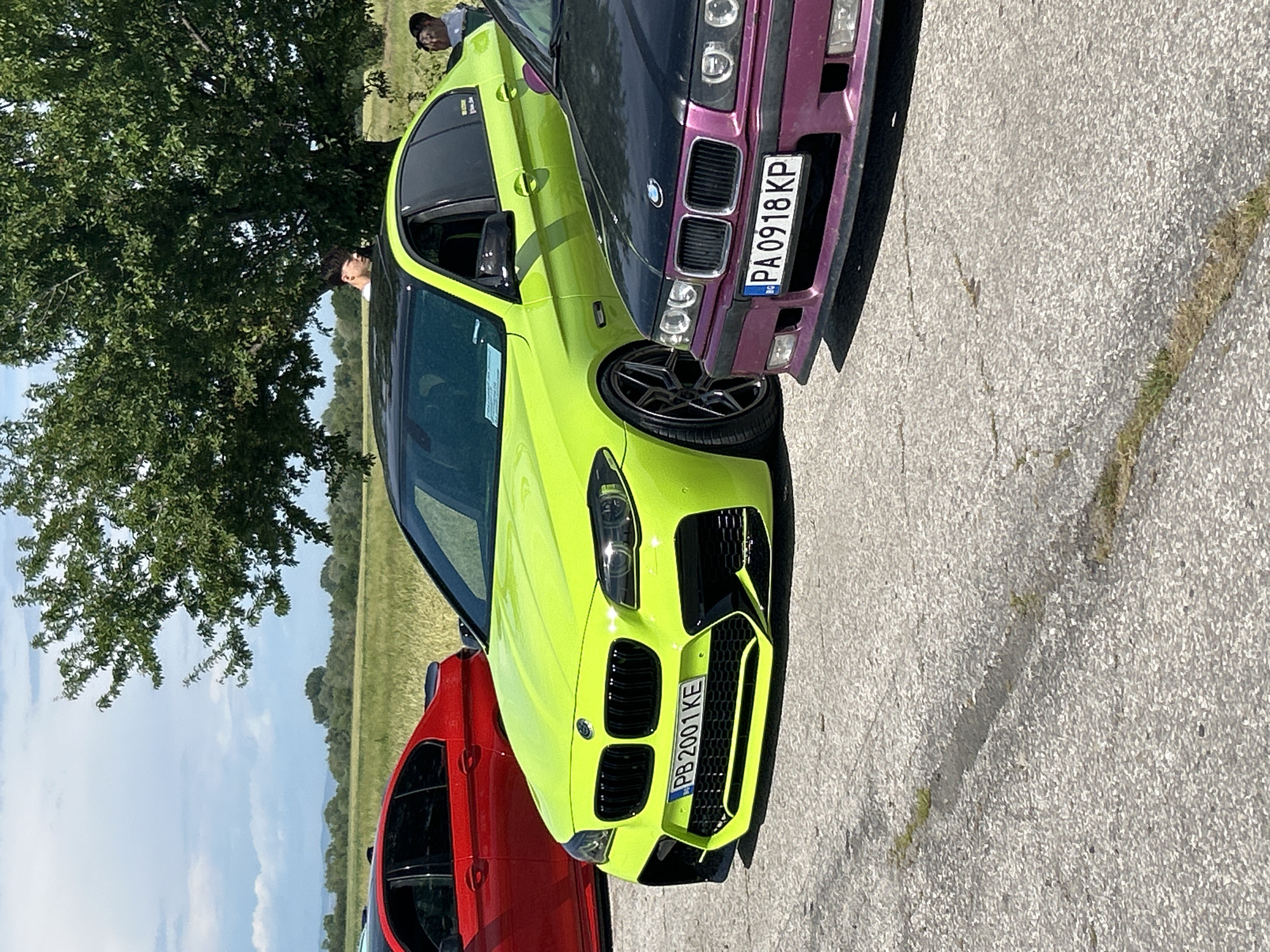 The image shows a car meetup on a concrete surface. A neon green BMW is in the foreground, with a Bulgarian license plate "PB 2001 KE". To the left is a red car, and to the right is a purple BMW, license plate "PA 0918 KP". A tree is in the background, and a field is visible in the distance. It is daytime, and the lighting is bright. A person is visible behind the cars. This photo was taken in Dalbok Izvor, Bulgaria.
