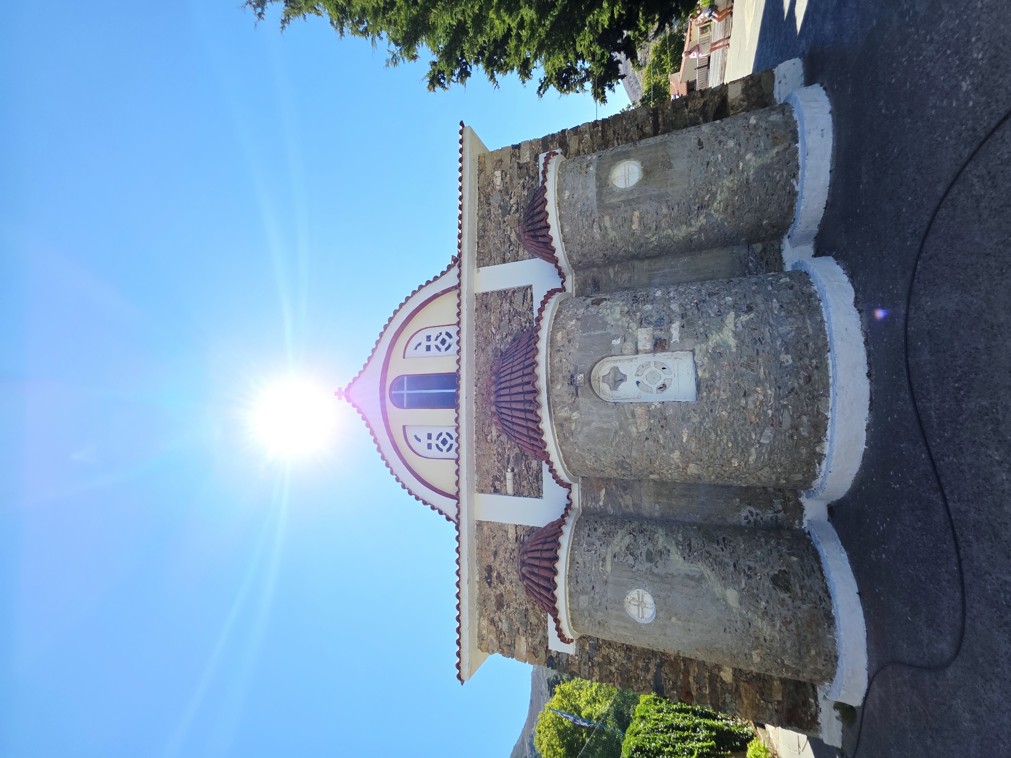 The image shows a stone church with multiple rounded structures on its facade, topped with red tiled roofs. The church has white trim around the roofline and around the rounded sections. Above the entrance, there is a semi-circular arched window with a cross design within it. The sun is shining brightly from the upper center of the frame, creating lens flare and casting strong light on the scene. The church is set against a clear blue sky. To the left of the church, there is lush green foliage and a blue and white flag. To the right, a tall green conifer tree is visible, and in the background, there are buildings and a road with two women standing. The foreground shows a dark asphalt surface. The image appears to be taken in Elos, Greece, a location known for its traditional stone architecture.