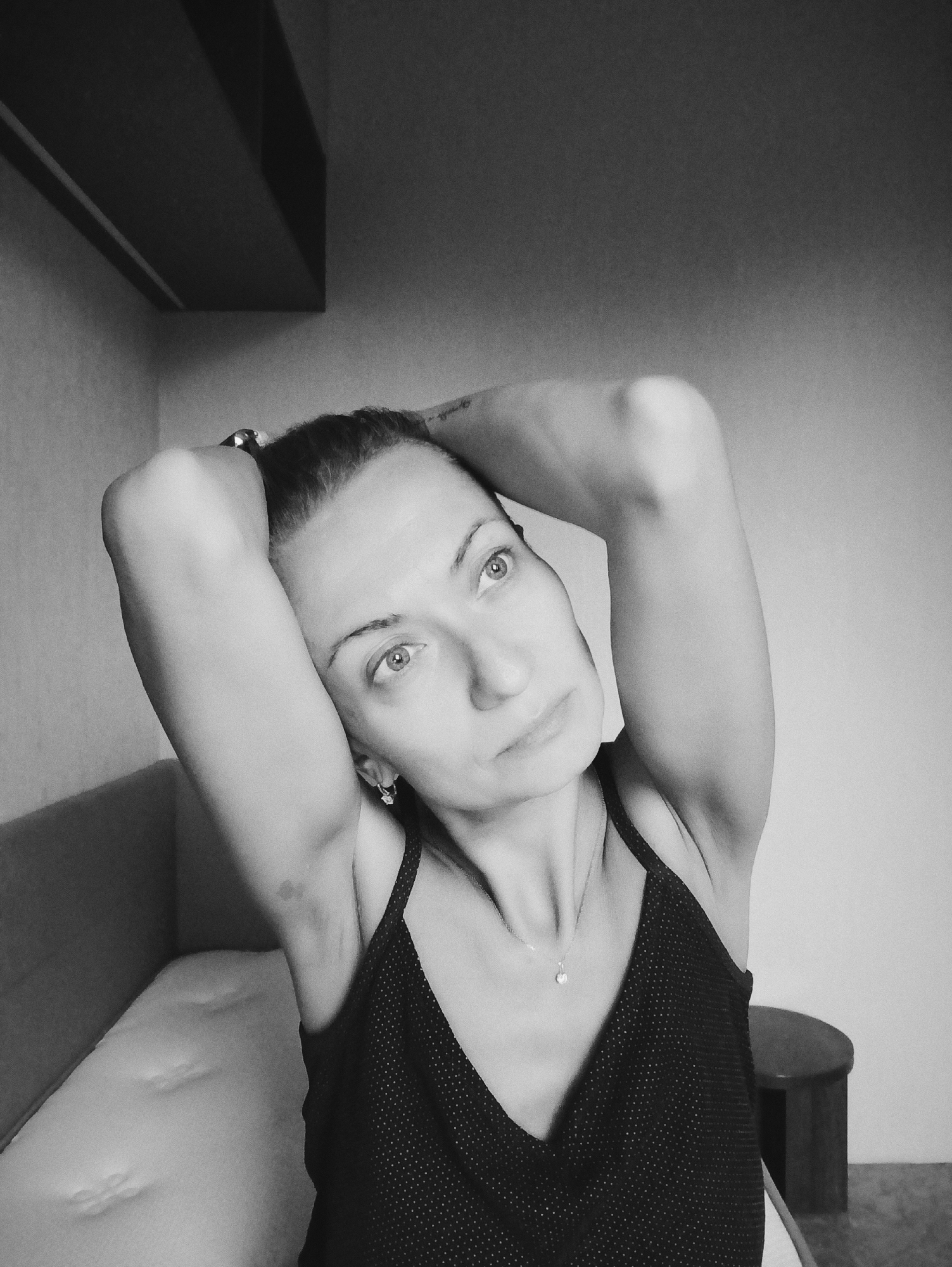A black and white medium shot shows a woman with her hands behind her head, looking up and to the right. She is wearing a sleeveless, polka-dotted top and a delicate necklace. Her expression is contemplative. The background is dimly lit and appears to be an indoor setting, possibly a bedroom or living room, with a bed or couch visible on the left. A small stool is present on the right. The overall ambiance is intimate and serene. The specific location within Moscow, Russia, cannot be determined from the visual information provided in the image.
