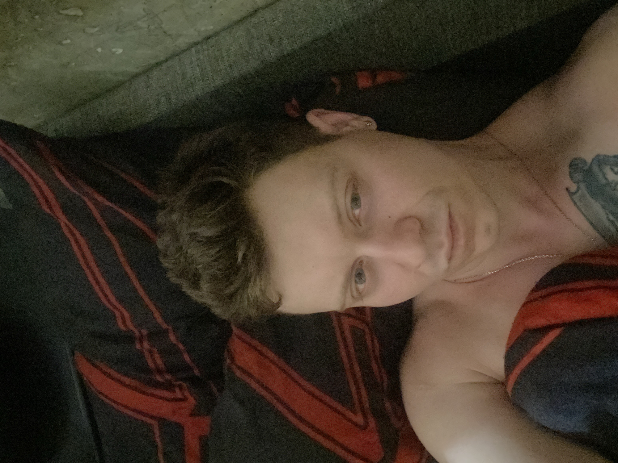 A close-up, high-angle selfie features a person lying down, partially covered by a dark blanket with red stripes. The person has short, brown hair and is looking towards the camera with light blue eyes. A faint smile is visible on their lips. They are wearing a thin silver chain around their neck and a small stud earring in their visible ear. A tattoo is partially visible on their chest. The background consists of a textured surface that appears to be a sofa or bed. The lighting is dim, suggesting an indoor setting, possibly a bedroom. The scene is devoid of any identifiable landmarks or characteristics that would indicate a specific location within Samara, Russia.
